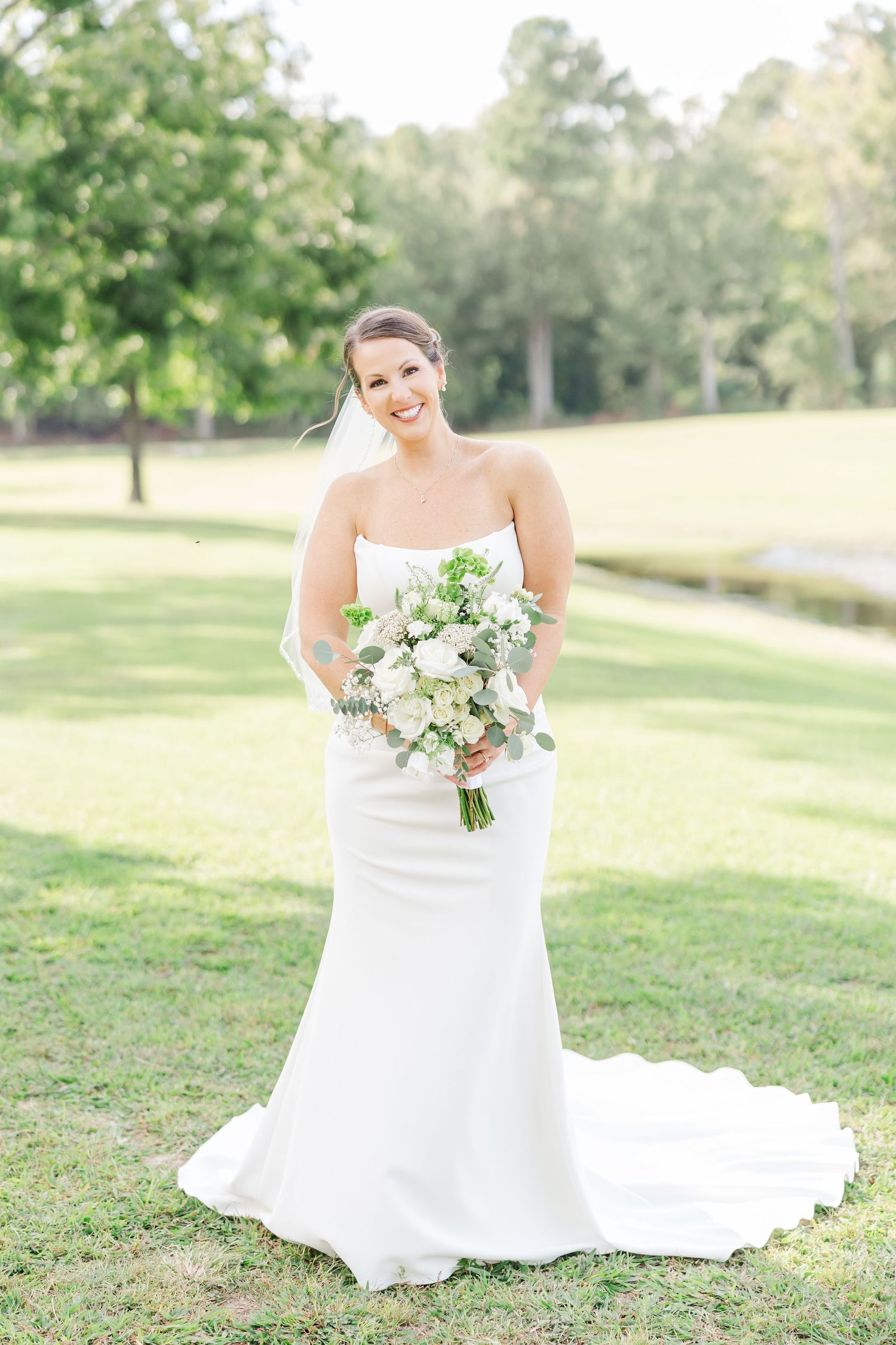 A bride in a flowing white wedding dress poses gracefully on a sunlit lawn holding a delicate bouquet of white flowers.