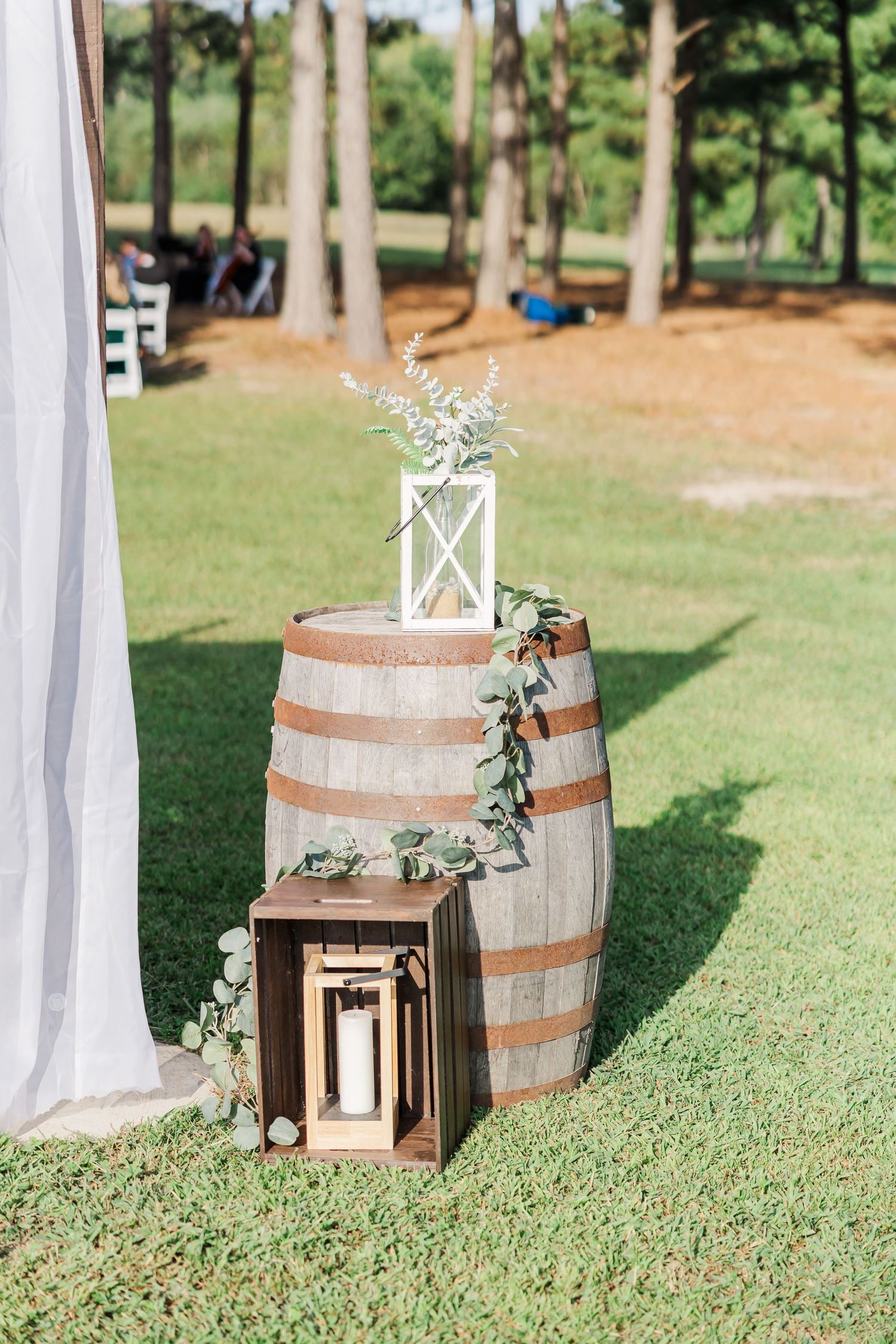 Rustic wedding decor with wine barrel, white lantern, greenery, and wooden crate on lush grass outdoors.