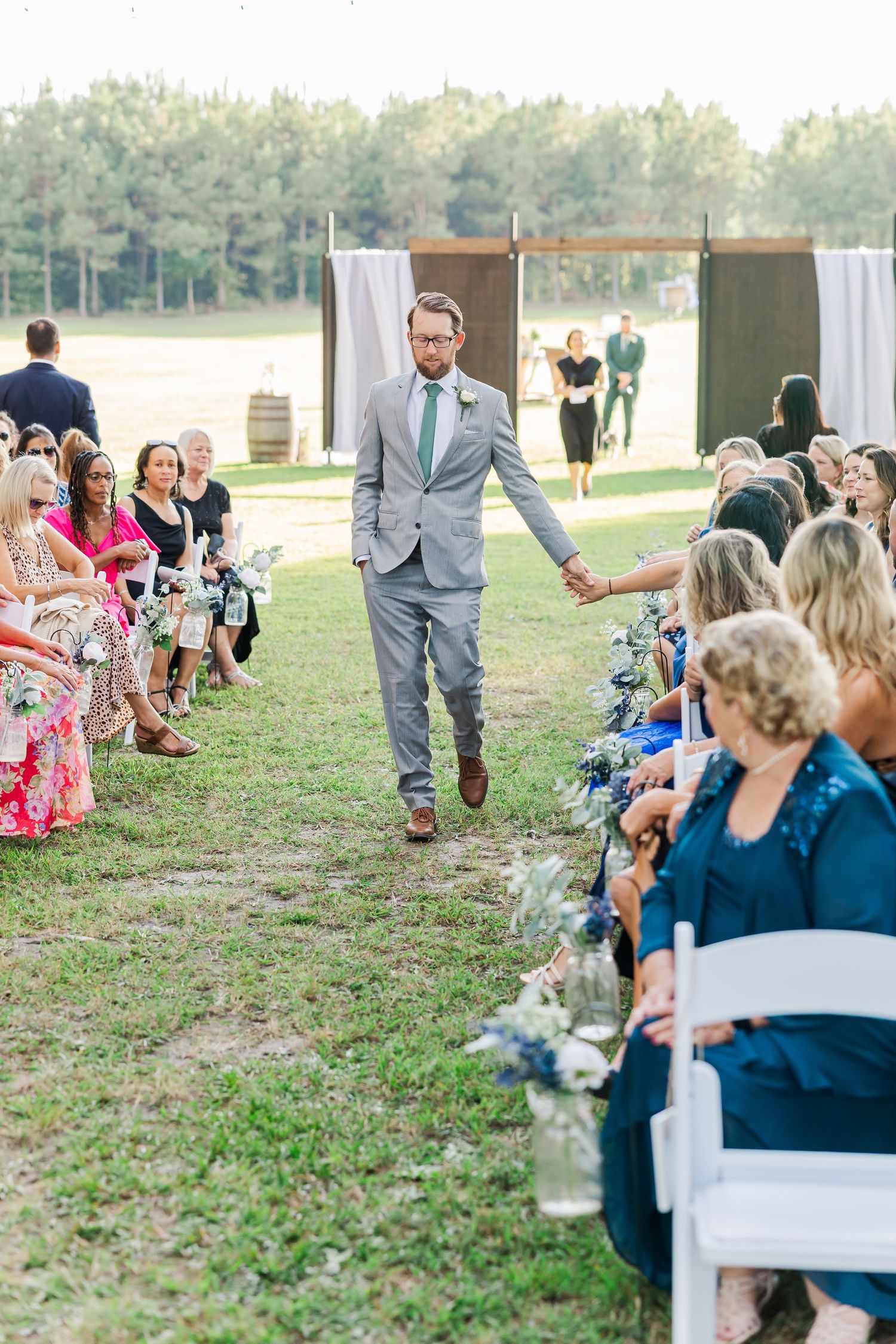 Groom in gray suit walks down outdoor wedding aisle holding guests hands with seated attendees on both sides.