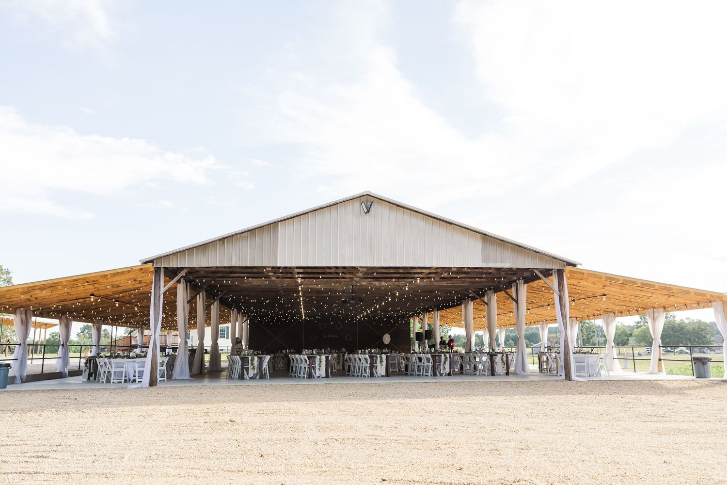 Large open-air barn wedding venue with string lights, reception tables, and a spacious sandy floor under a wide roof.