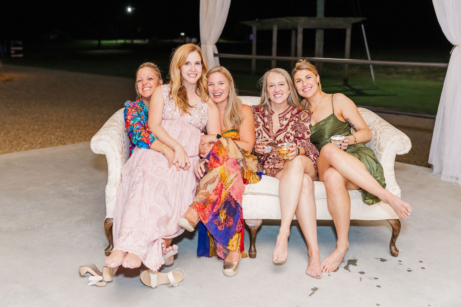 Five women in colorful dresses smiling together on a white sofa at an outdoor evening event.