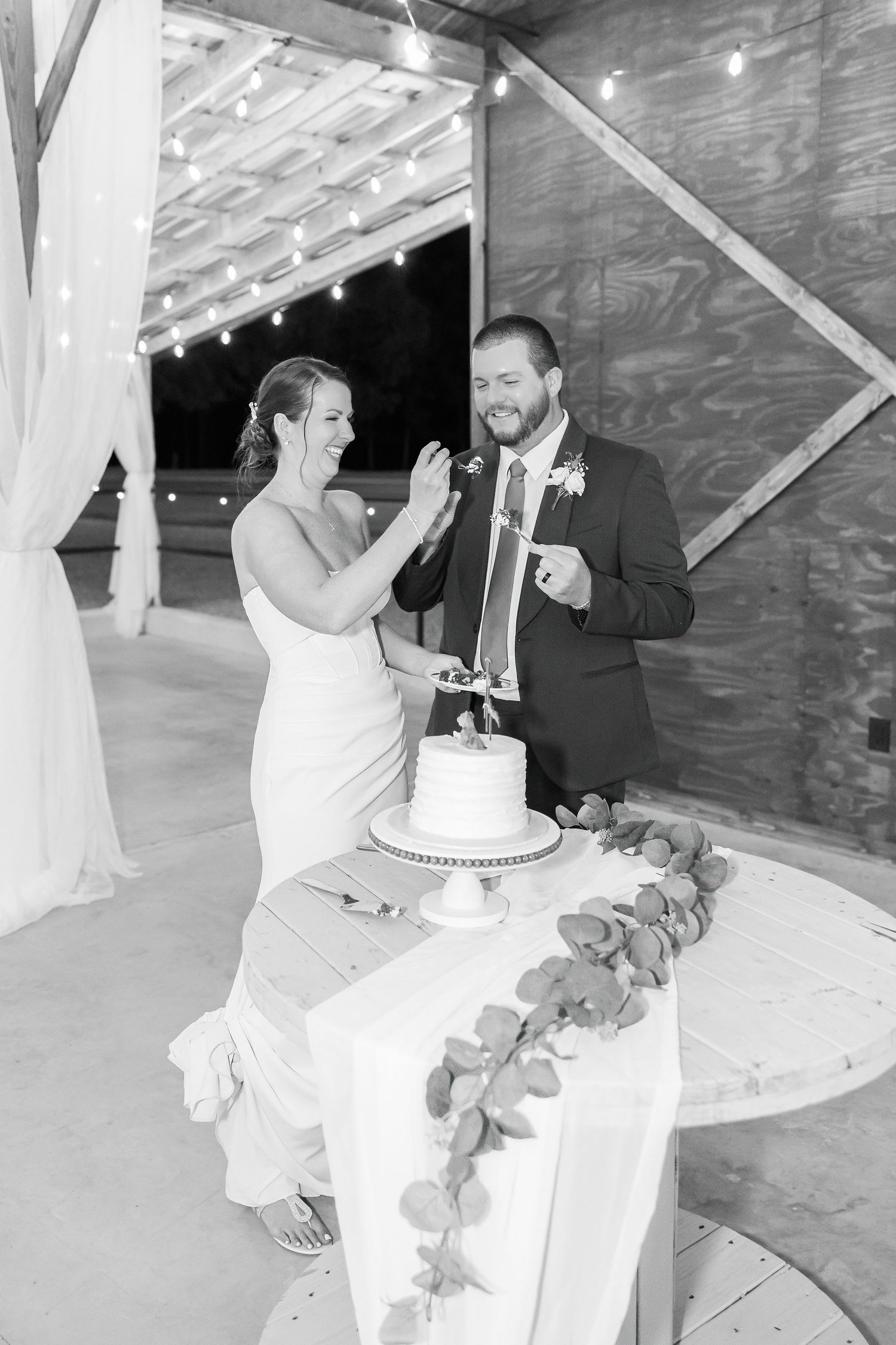 Black and white photo of bride and groom laughing while cutting wedding cake at rustic barn venue with string lights.