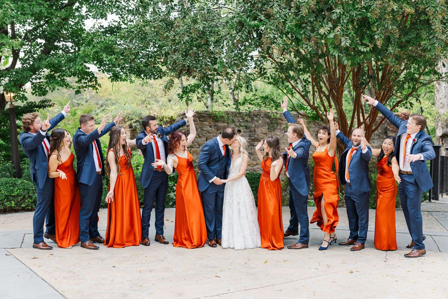 Wedding party in navy suits and bright orange bridesmaid dresses poses together outdoors under green trees.