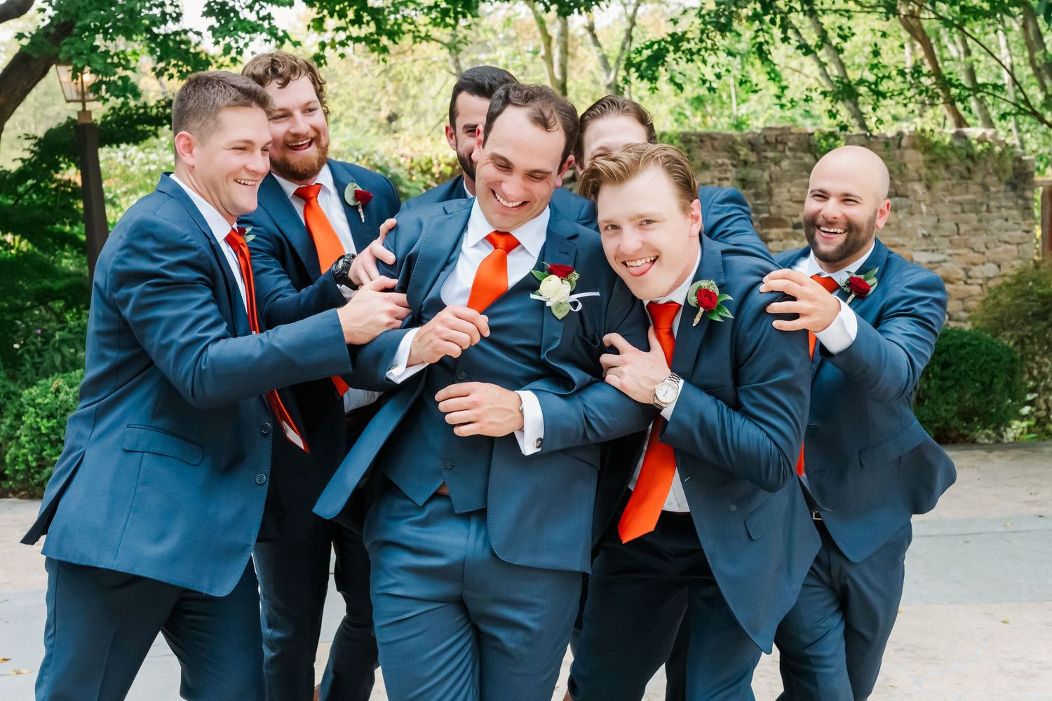 Wedding party in navy suits and orange ties laugh together while posing for photos outdoors.
