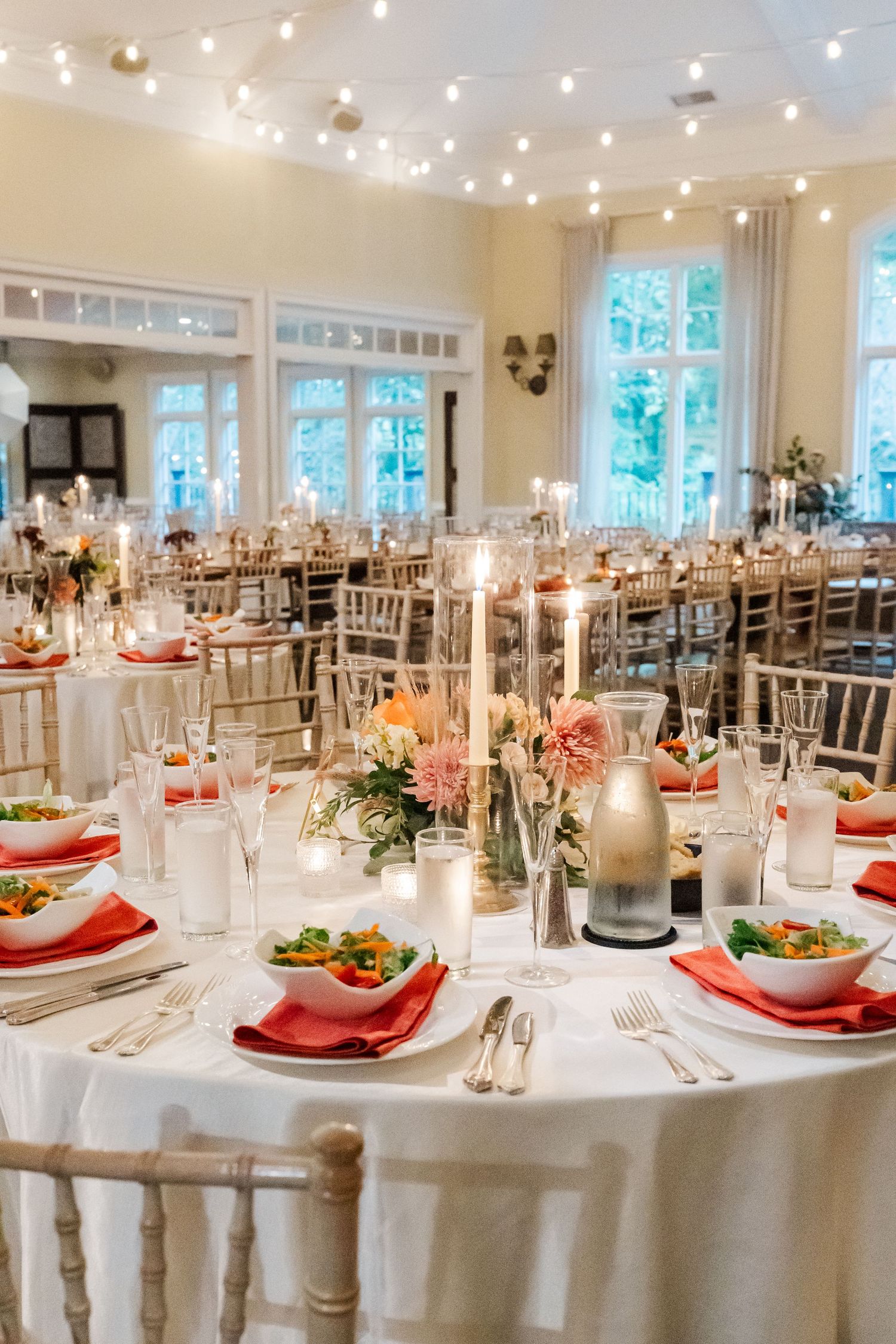 Elegant wedding reception tables set with red accents, white linens, and twinkling overhead lights.