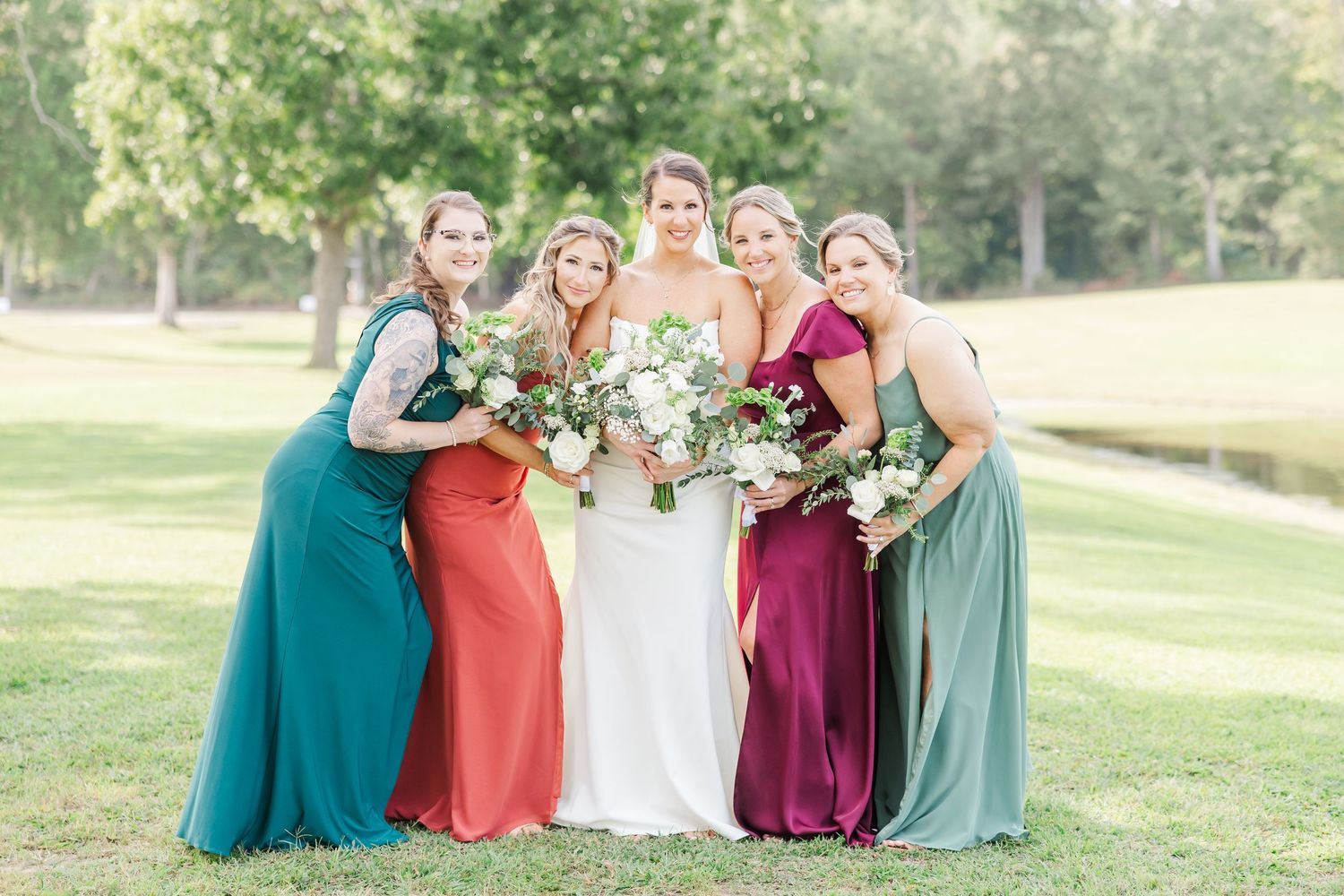 Bridal party in jewel-toned dresses poses together holding bouquets on a grassy lawn.