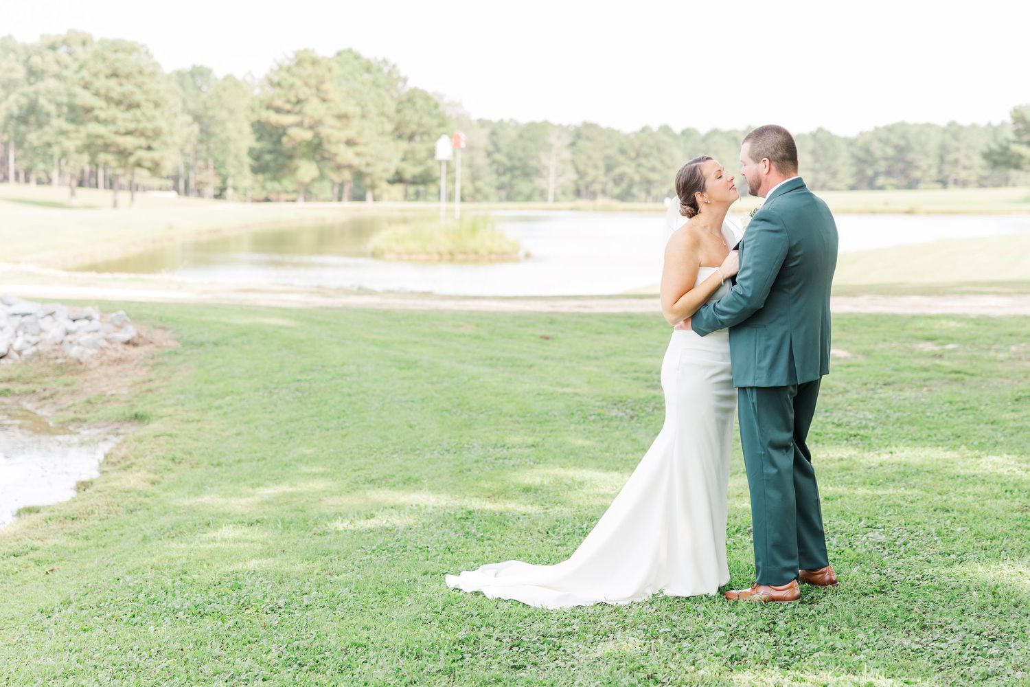 A couple shares a romantic moment in their wedding attire on a grassy lawn near a scenic pond.