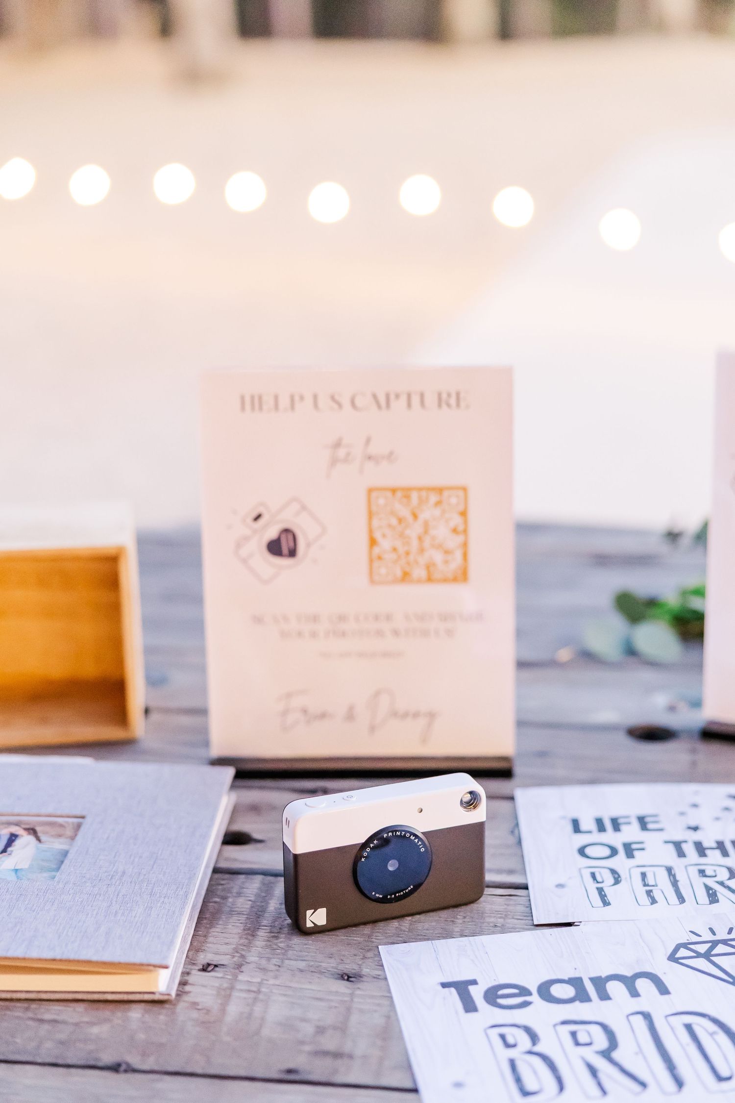 A Kodak instant camera sits beside a QR code guest photo sign and Team Bride cards on a rustic wooden table at a wedding.