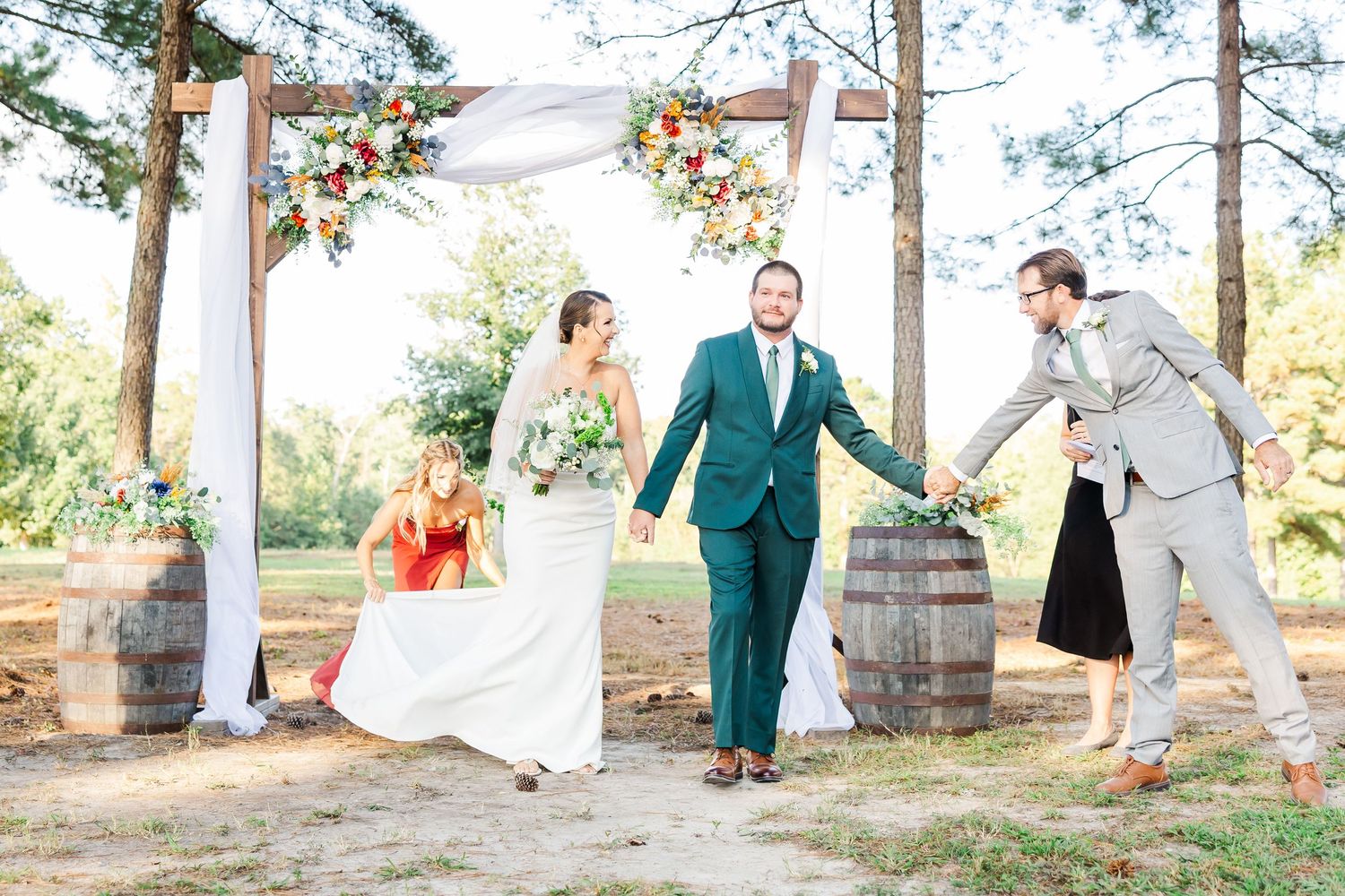 Joyful newlyweds celebrate under a floral arch with wedding party at an outdoor rustic ceremony.