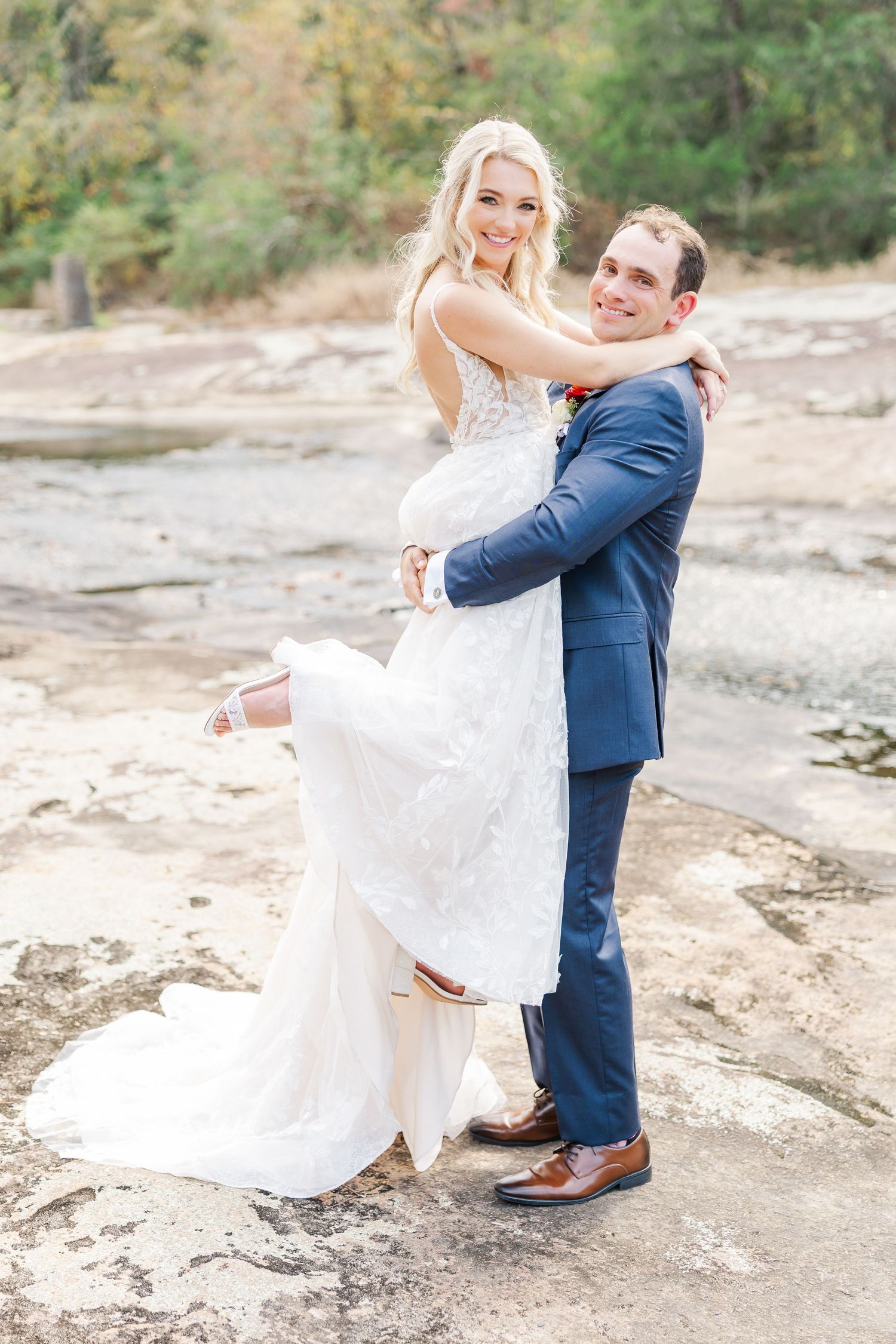 A joyful wedding couple embracing on rocky terrain with the groom lifting the bride in her lace gown.