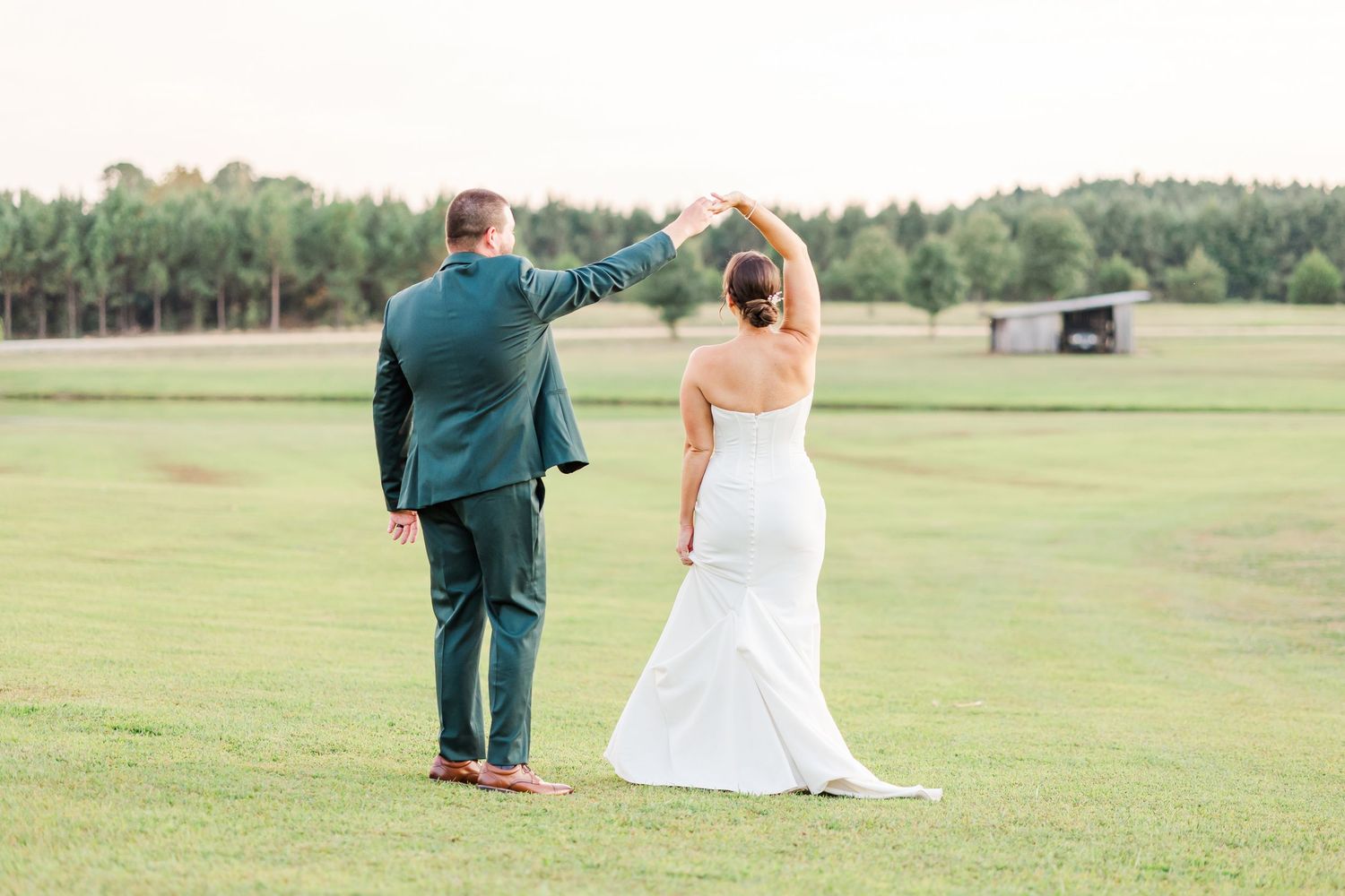A couple in wedding attire walks hand in hand across a lush green field on their special day.