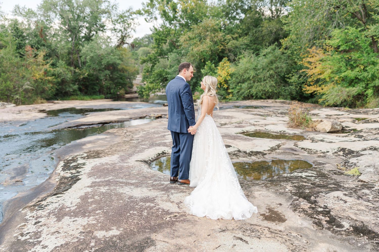 Newlyweds walk along scenic rocky riverbank with trees in the background.