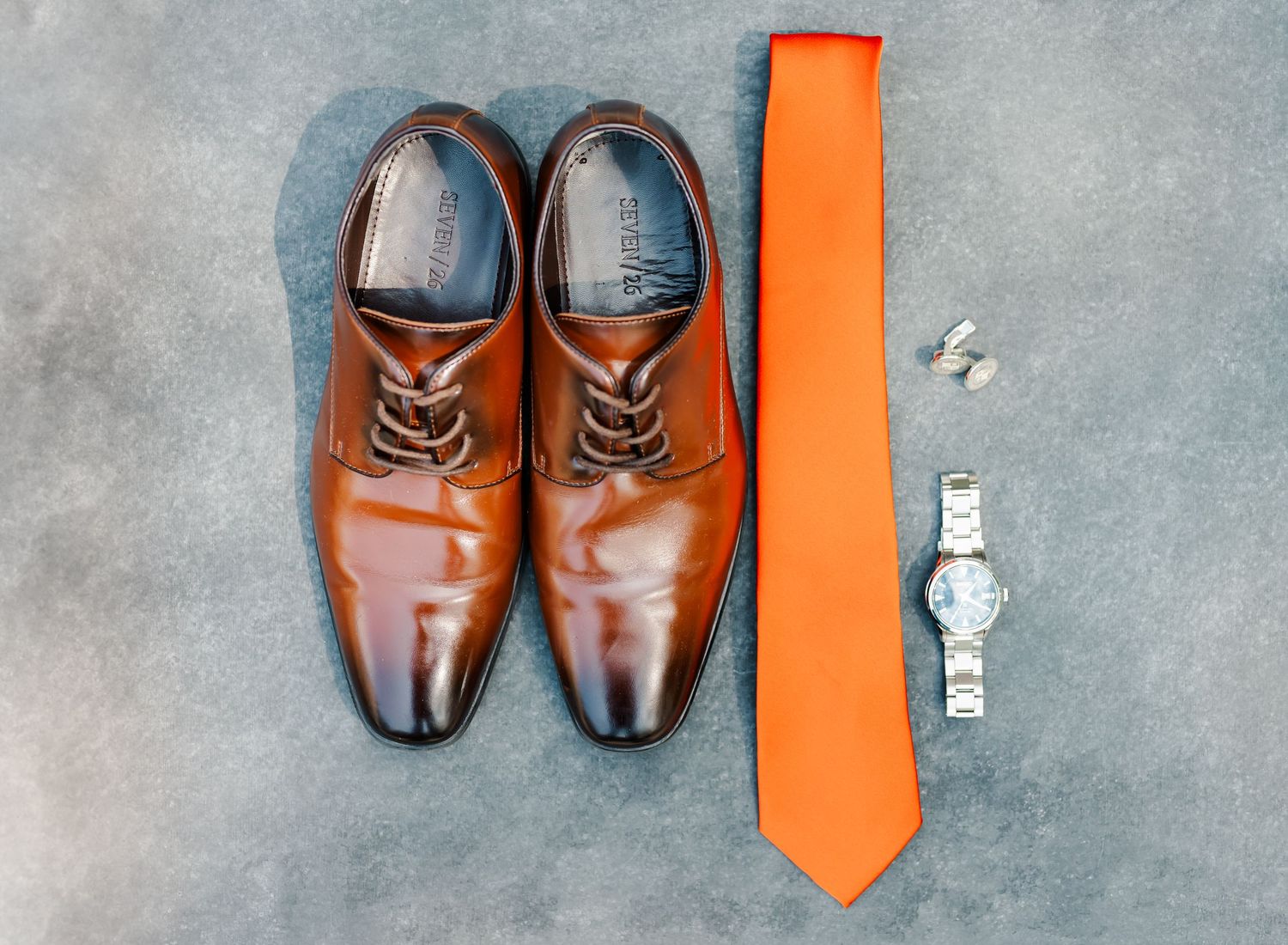 A collection of brown leather dress shoes and an orange necktie arranged in a row on a gray concrete surface.