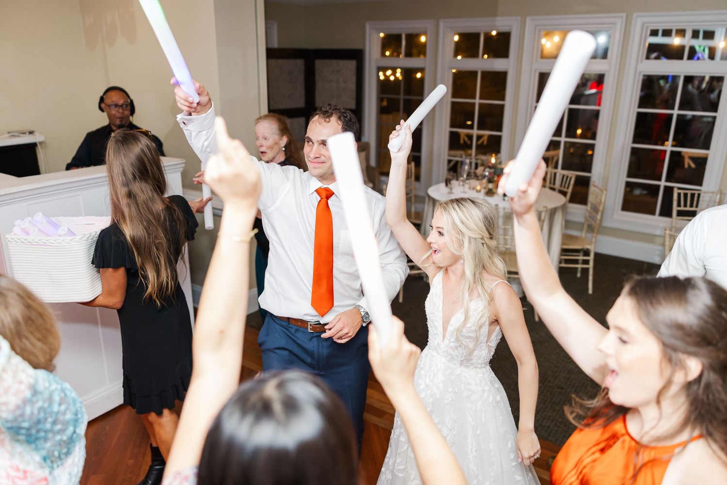 Wedding guests raise their hands while dancing at a lively indoor reception.