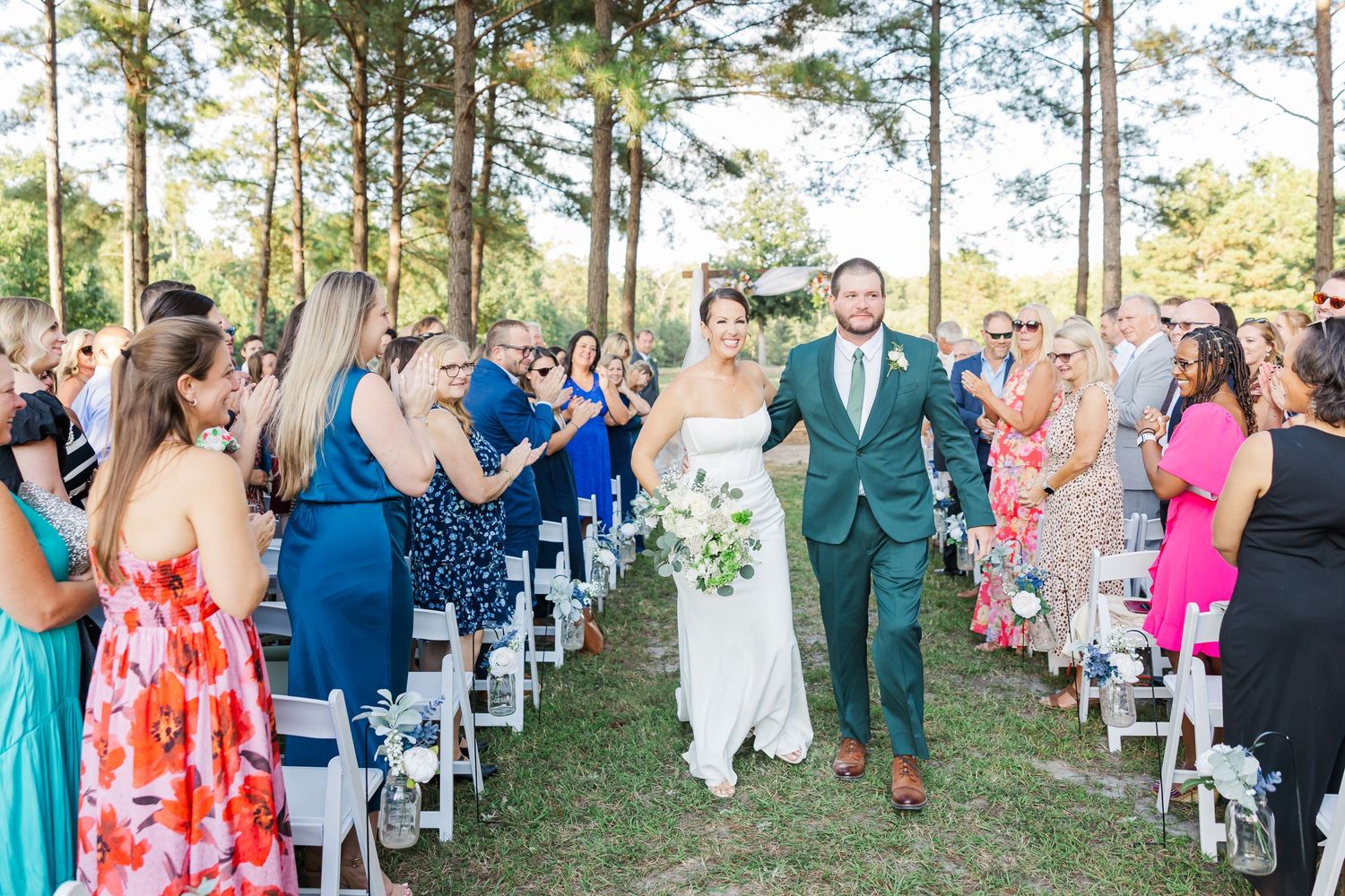 Newlywed couple walks back down the aisle after outdoor wedding ceremony as guests cheer and applaud.