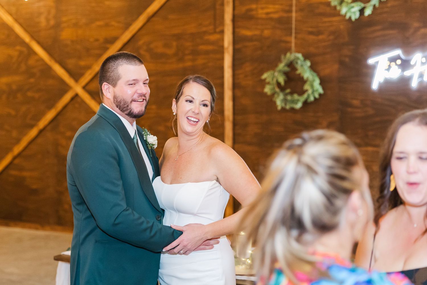 Smiling bride in white strapless gown and groom in teal suit stand together during wedding reception in rustic barn venue.