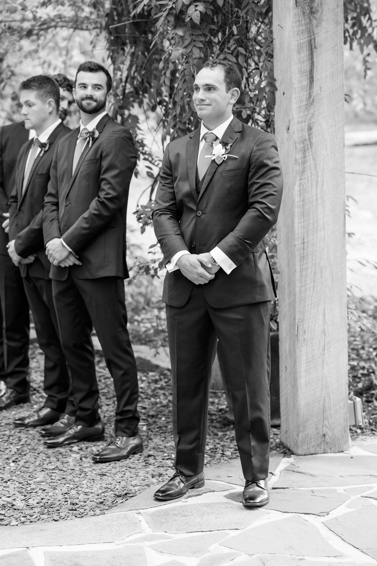 Groomsmen in dark suits stand together at an outdoor wedding ceremony location.