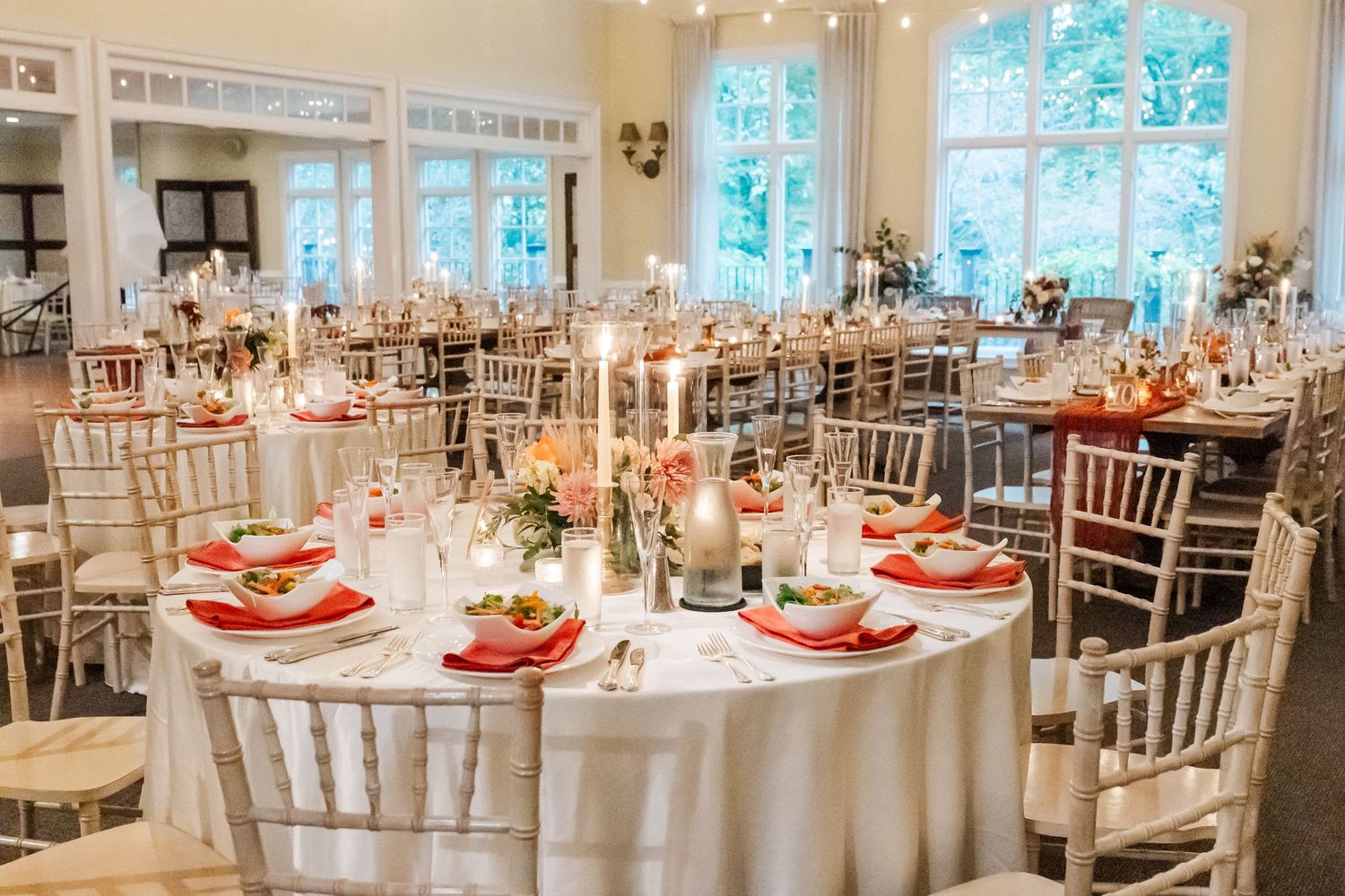 Elegant wedding reception venue with white chiavari chairs and red table accents in a bright, airy space.