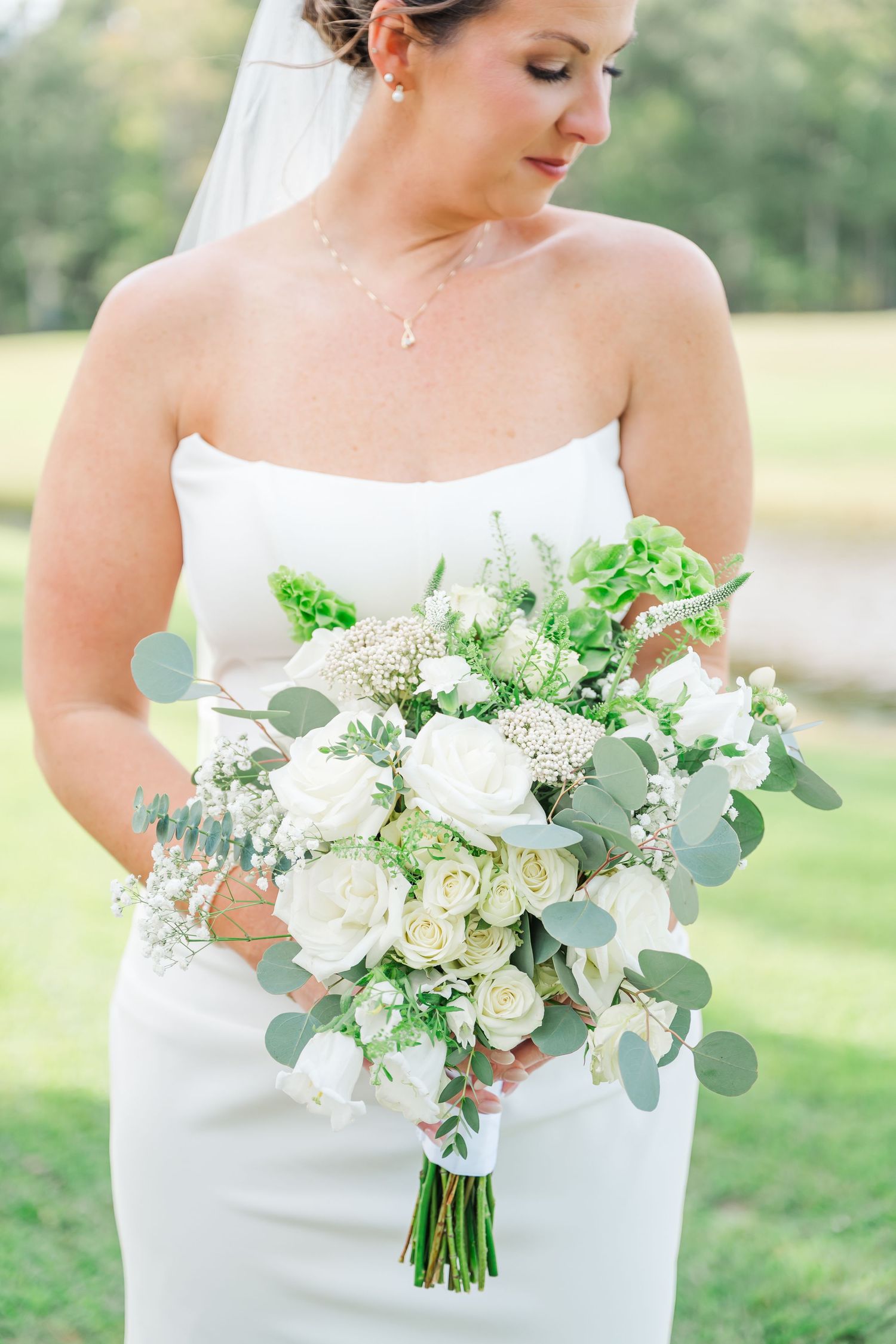 A bridal bouquet features white roses, eucalyptus leaves, and delicate green accents against a white dress.