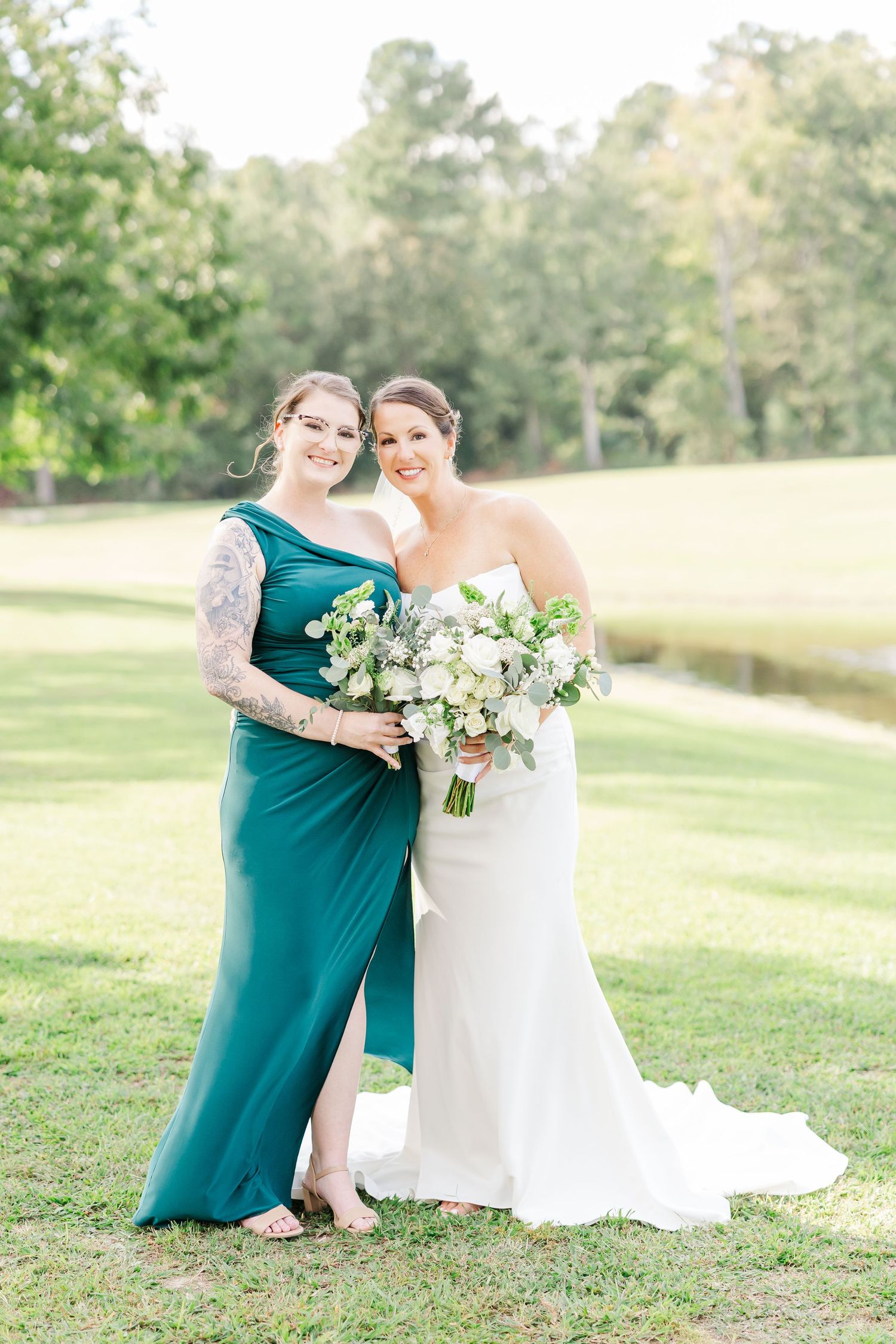 Wedding party members in teal dresses and white gown pose together on a sunlit grassy lawn with floral bouquets.