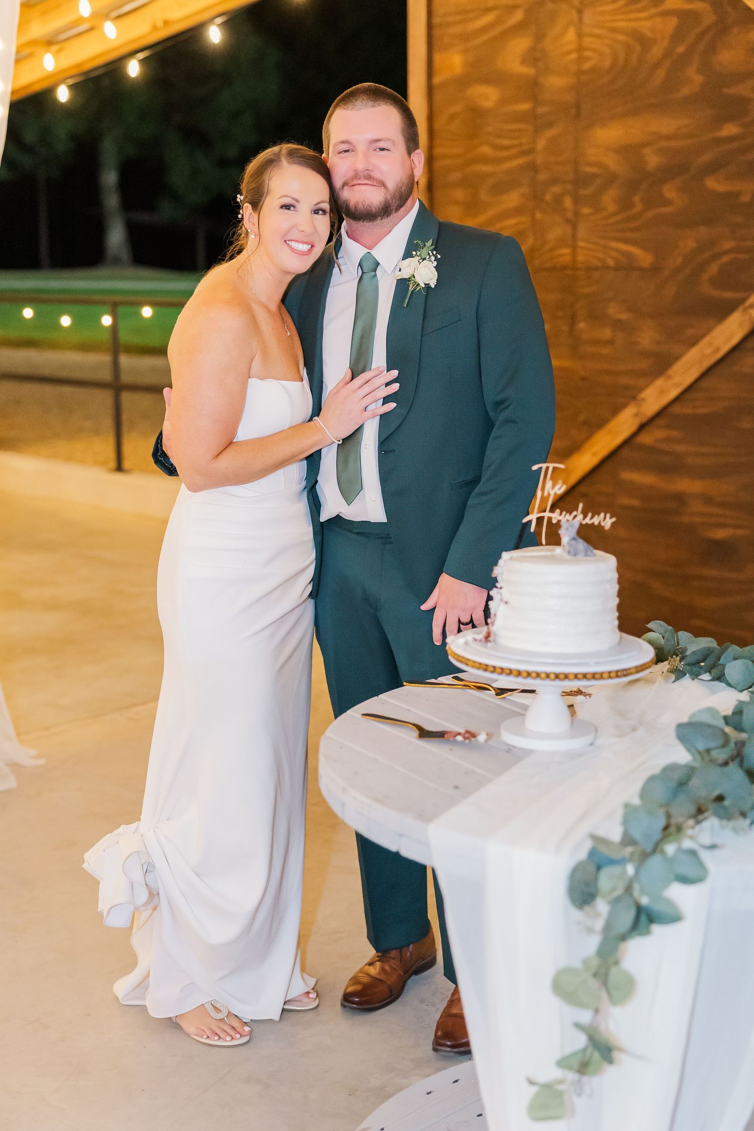 Bride and groom smiling beside white wedding cake with greenery, groom in teal suit, bride in white gown at outdoor reception.