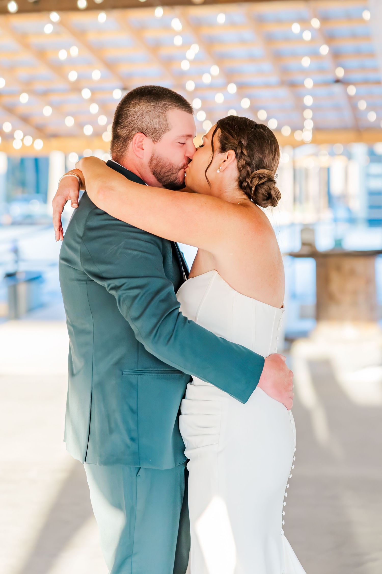 Bride and groom share a romantic first kiss in a teal suit and white strapless gown under twinkling string lights.