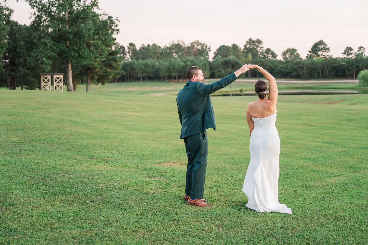 A couple dances together on a lush green lawn with trees in the background during a wedding celebration.