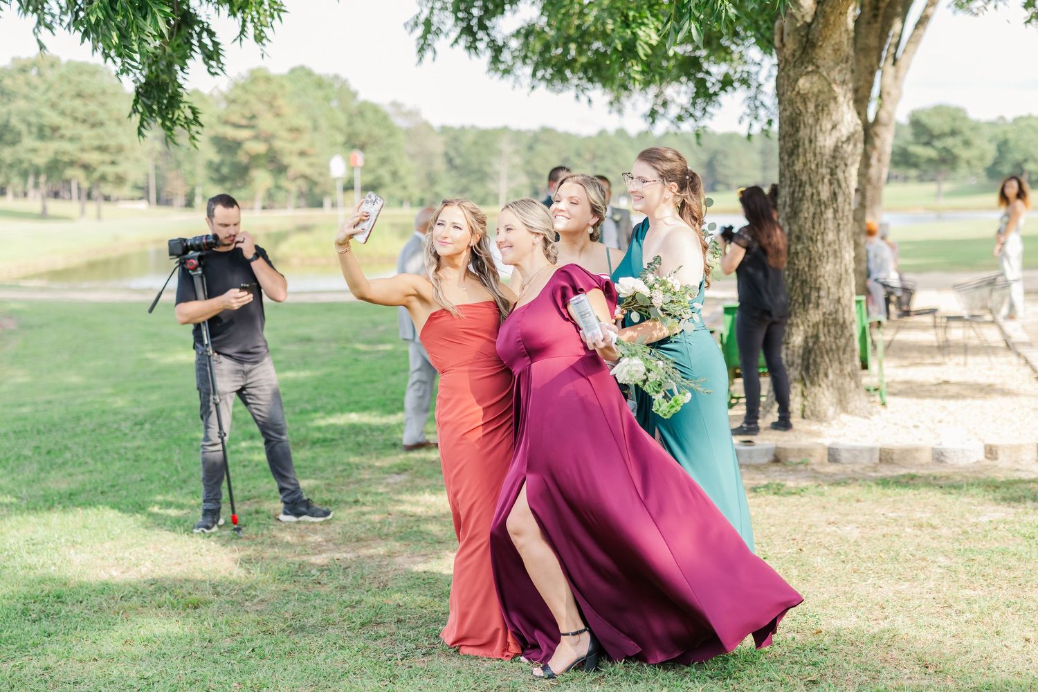 Wedding party in colorful dresses poses playfully in a scenic outdoor setting with trees and grass.