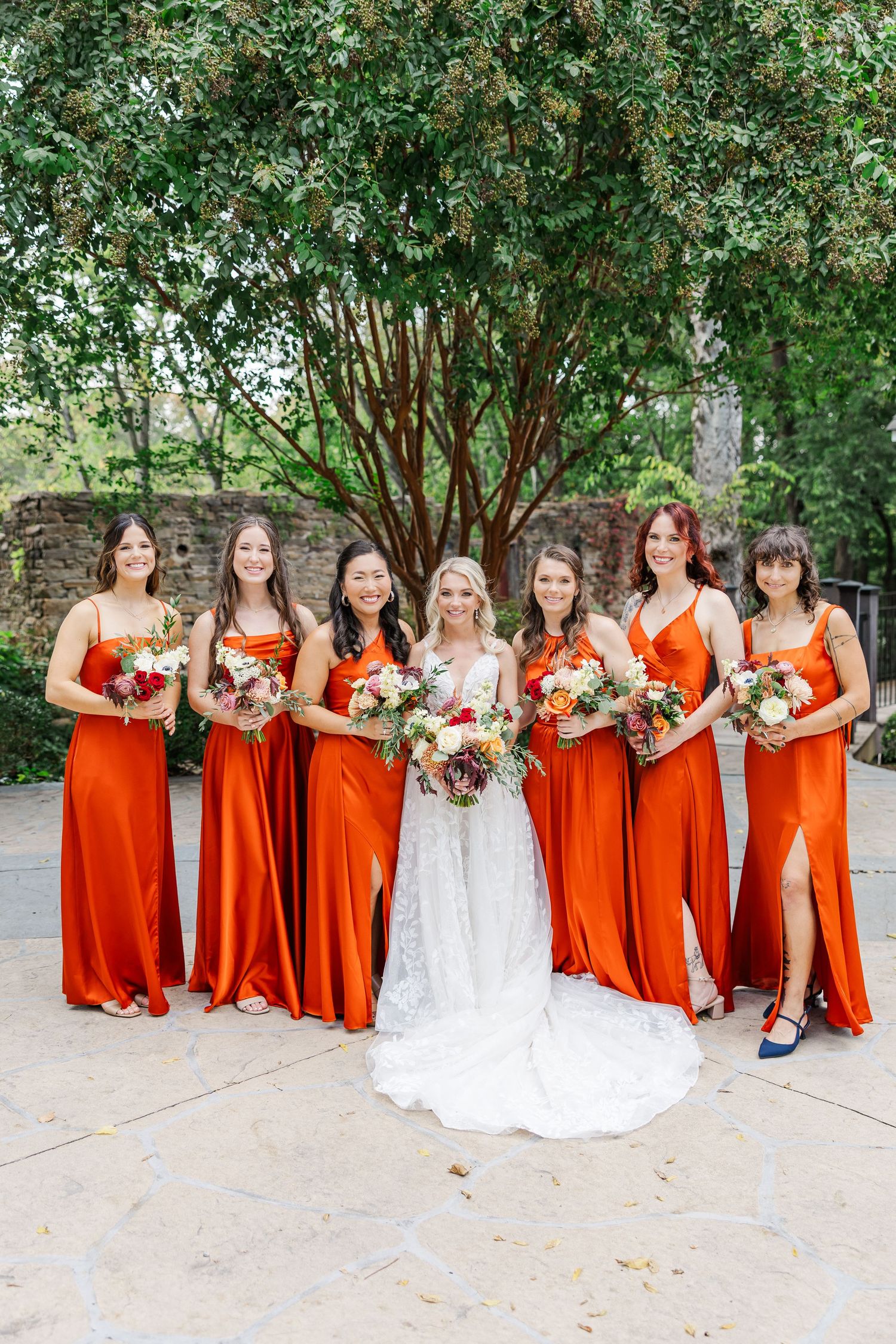 Bridal party in white wedding gown and vibrant orange dresses holding colorful bouquets outdoors.