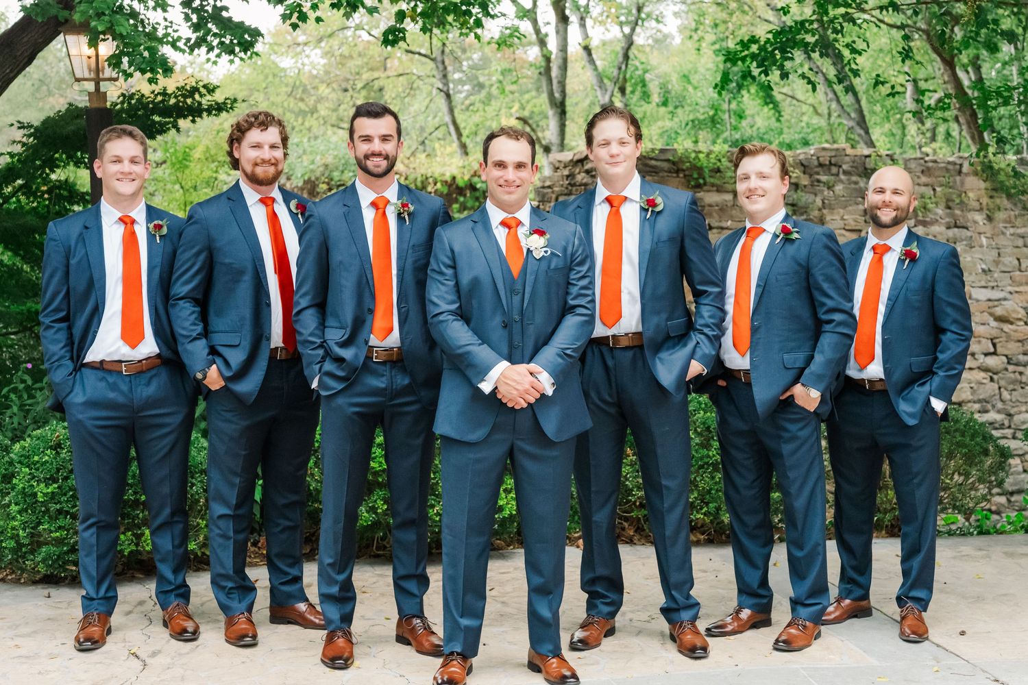 A wedding party in navy suits with bright orange ties and brown shoes pose together outdoors.