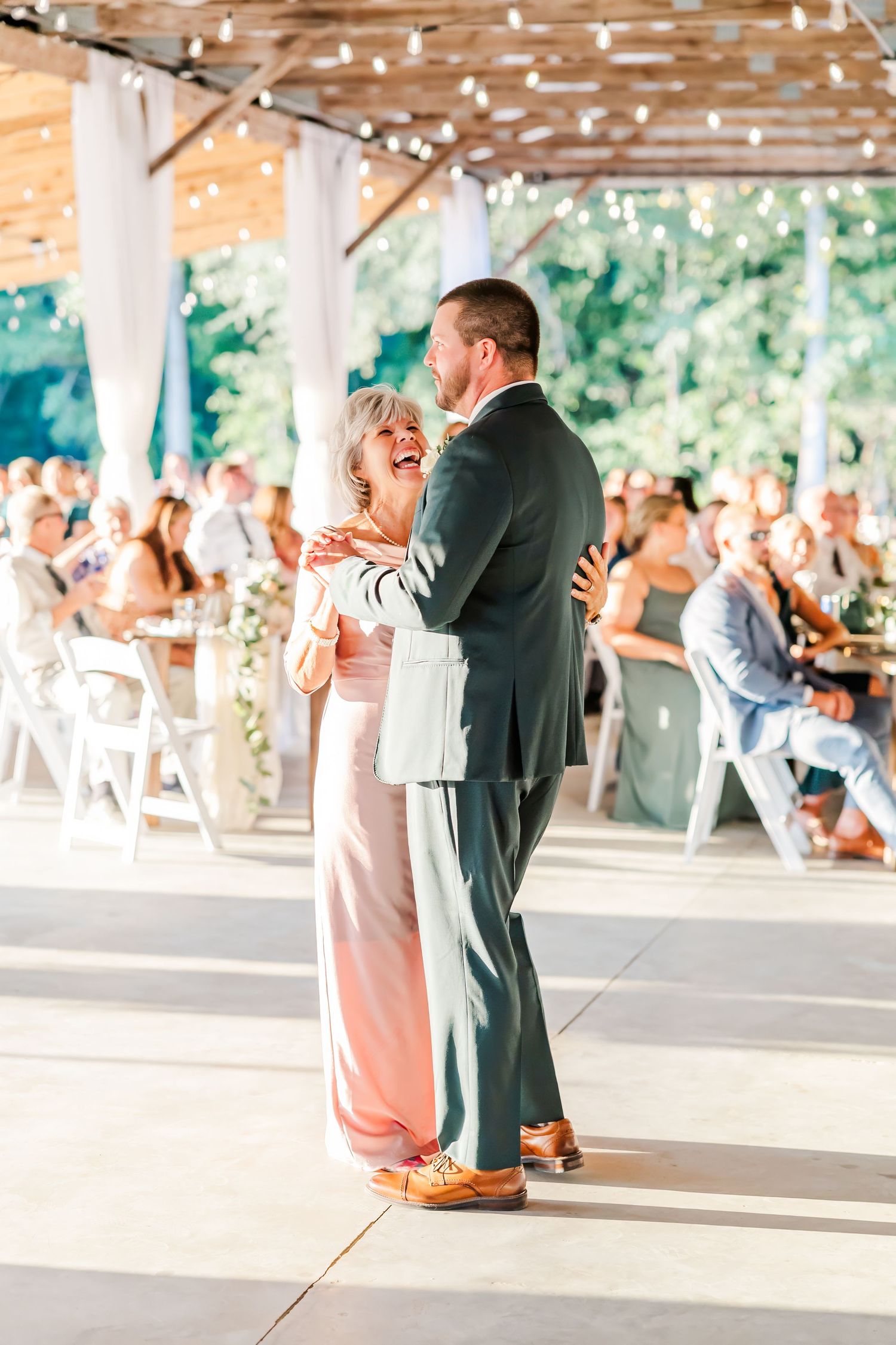 A groom shares a joyful mother-son dance at an elegant outdoor wedding reception with string lights and seated guests.