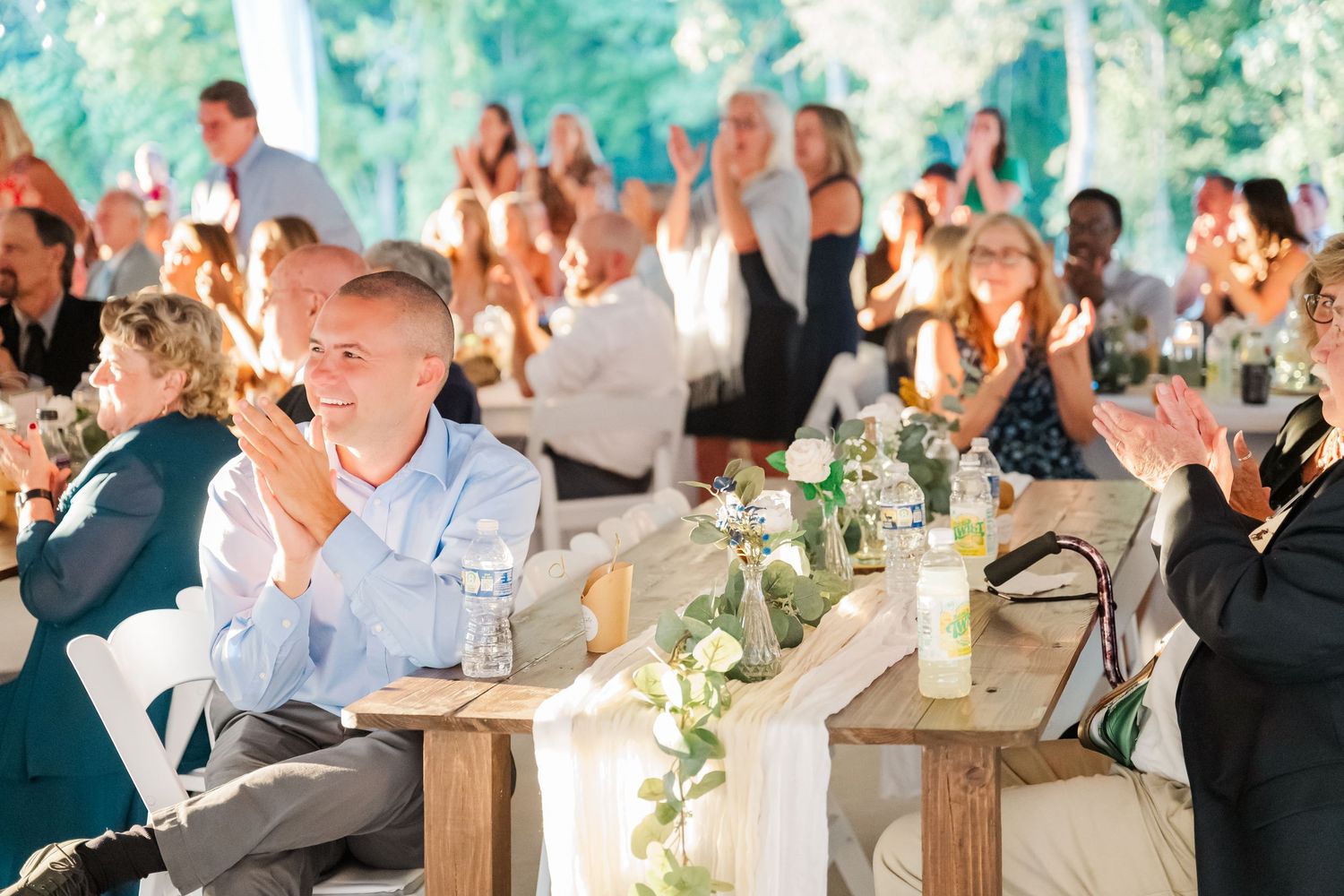 Wedding guests clap and celebrate at an elegant outdoor tented reception with floral centerpieces.