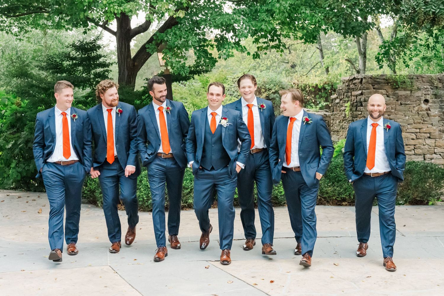 Wedding party members in coordinated navy suits and orange ties pose casually on outdoor stone steps.