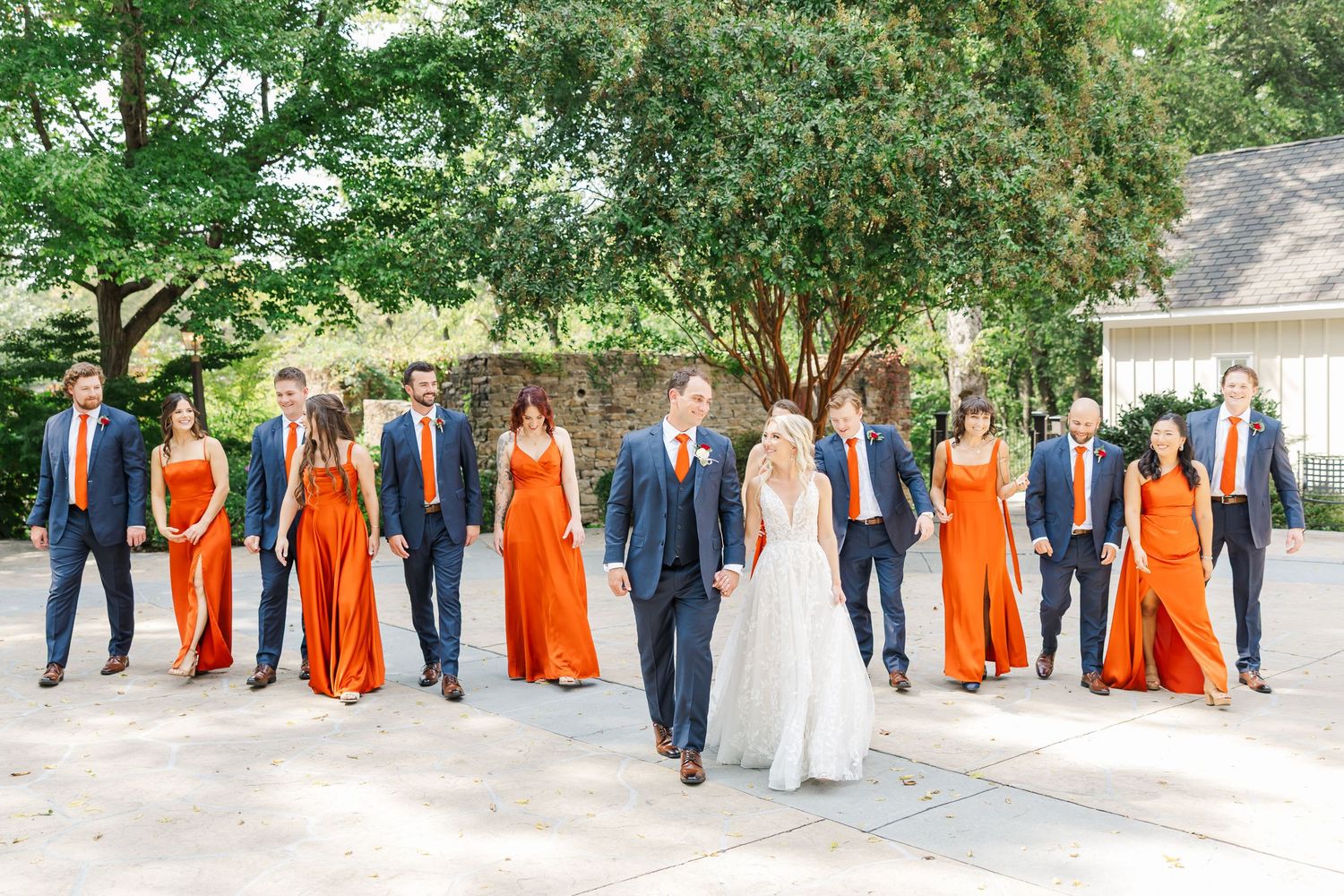 Large wedding party lineup with orange bridesmaid dresses and navy suits against natural outdoor backdrop.