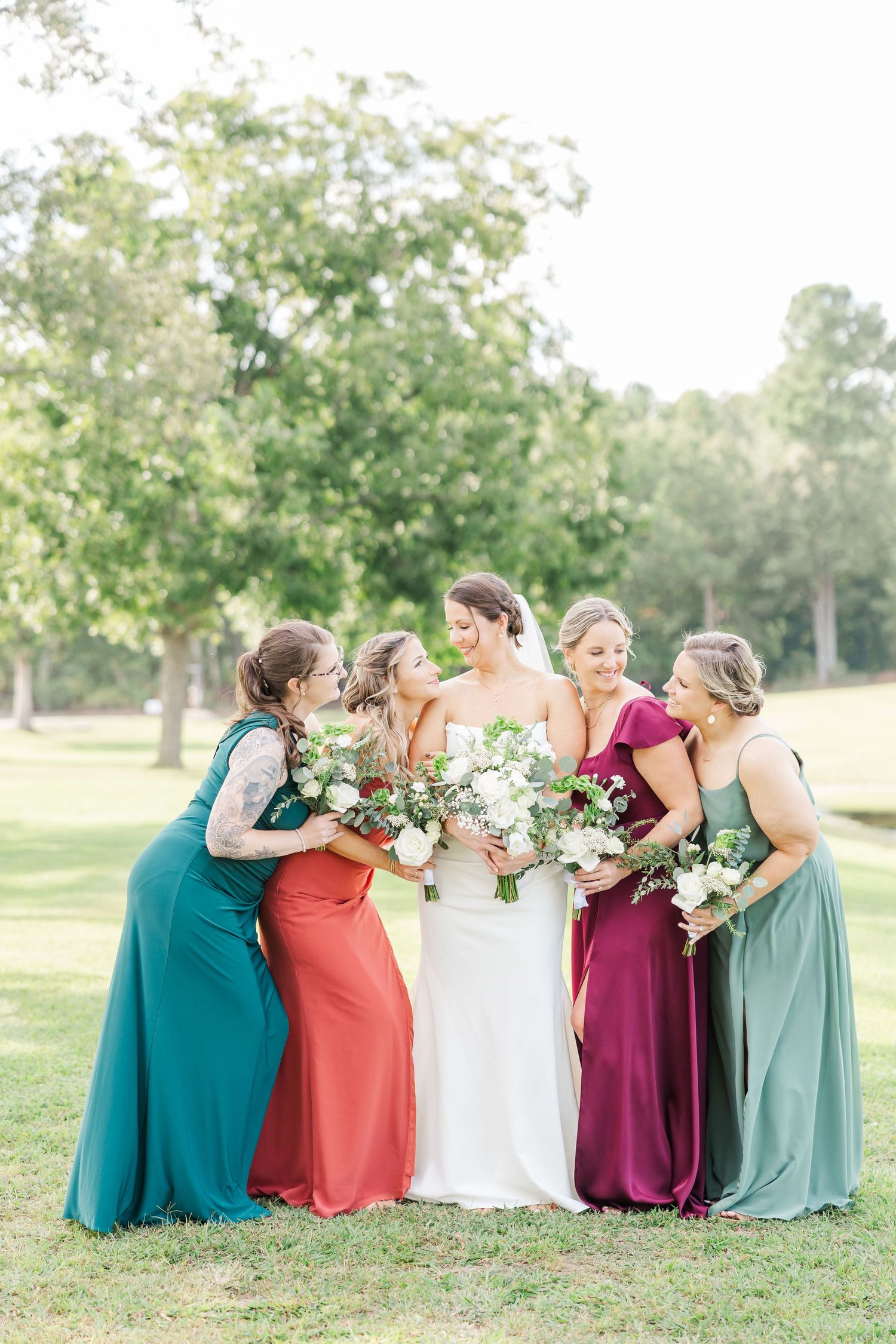 Bridal party in colorful floor-length gowns shares a joyful moment together outdoors.