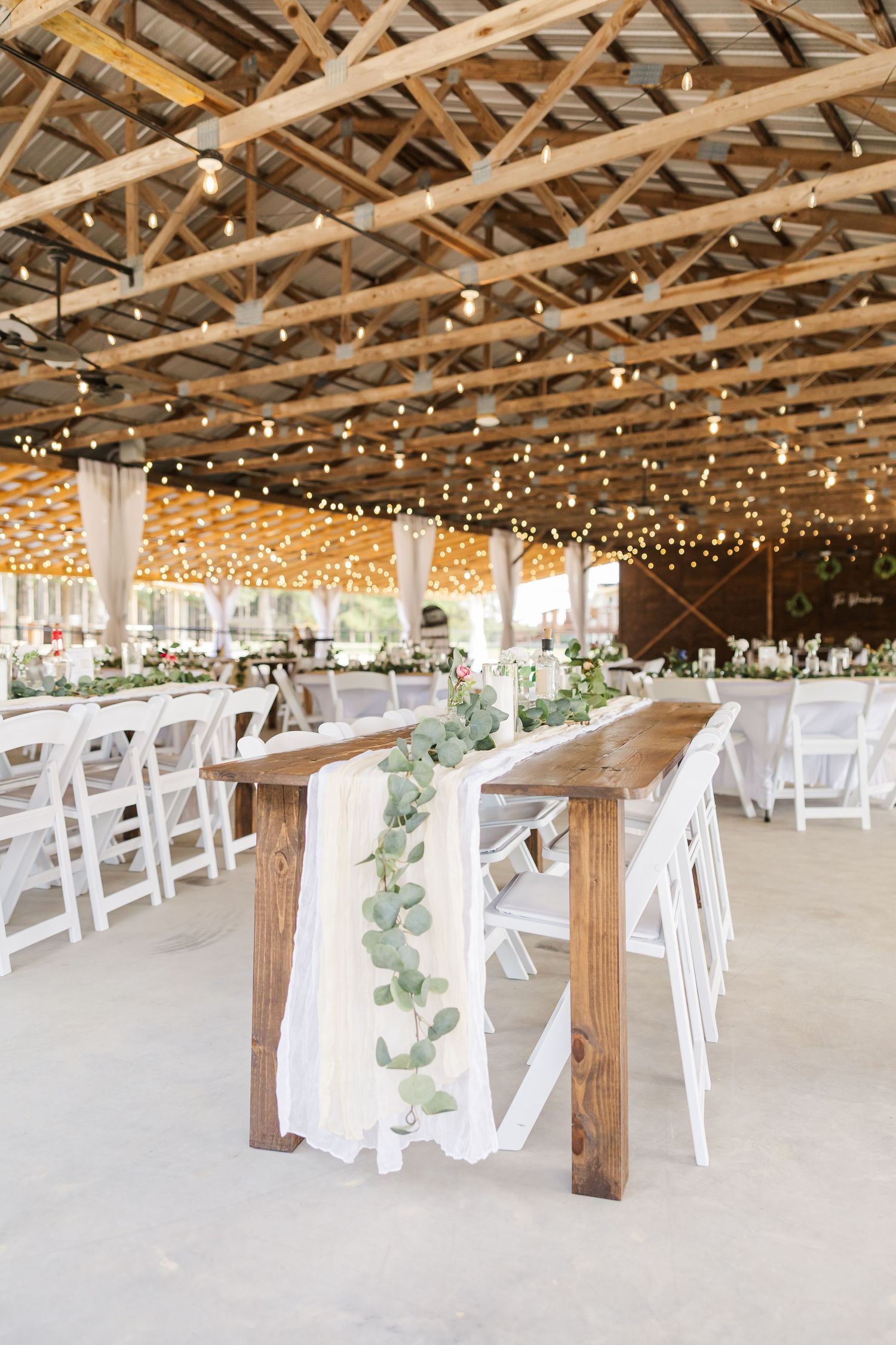 Elegant rustic wedding reception venue with wooden beams, string lights, white folding chairs, and eucalyptus table runners.