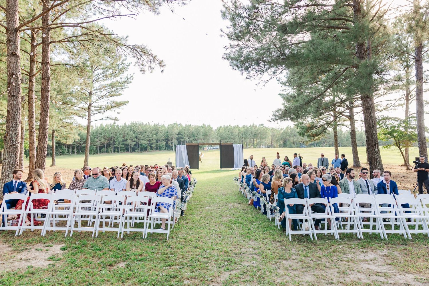 Outdoor wedding ceremony with rows of white chairs and guests seated among tall pine trees on a sunny day.