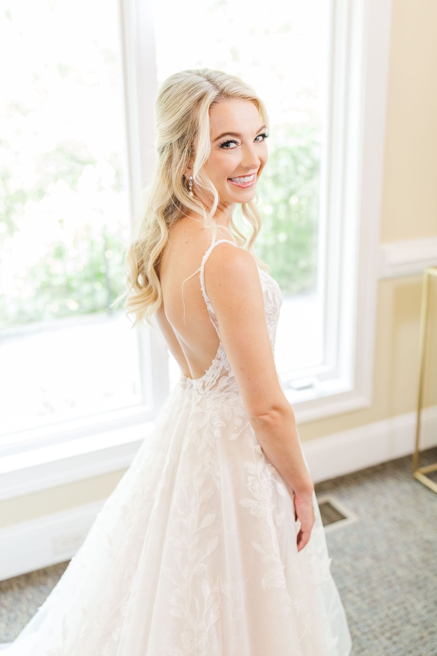 A bride in a flowing white dress stands by bright windows in an elegant room.