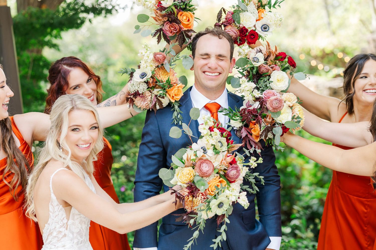 Wedding party poses outdoors in navy and rust orange attire with vibrant autumn floral bouquets.