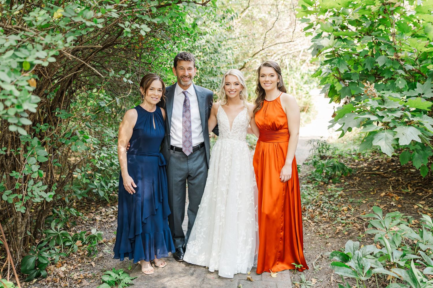 Wedding party in navy and orange dresses poses together outdoors in natural garden setting.