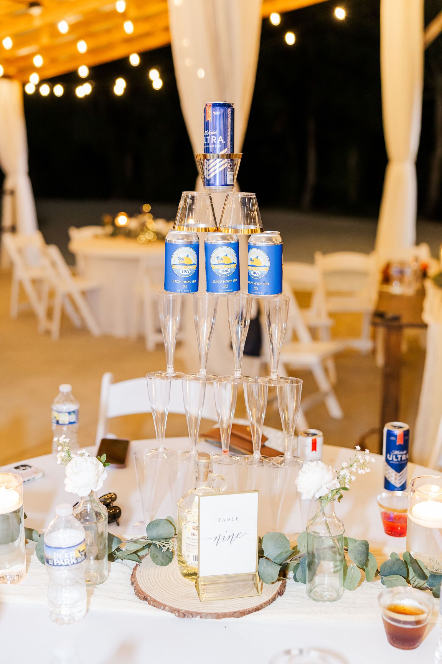 Elegant wedding reception beer can tower display with champagne flutes on wooden base, string lights, and floral decor.