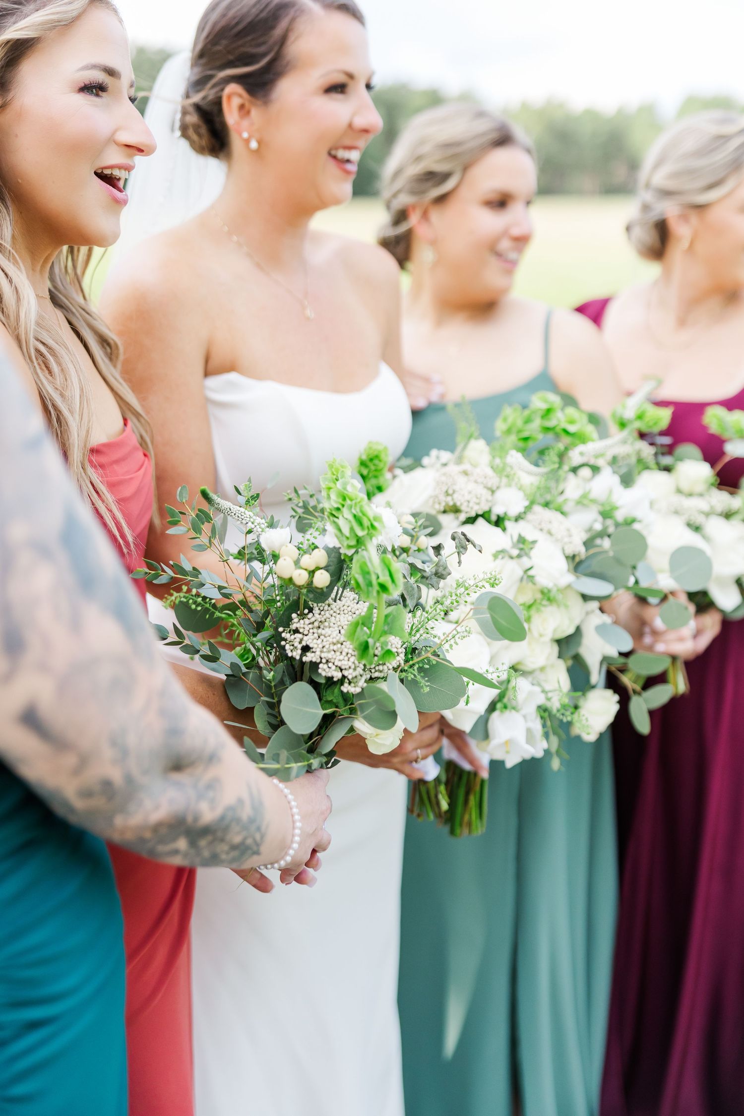 Bridesmaids in jewel-toned dresses holding elegant white and green bouquets at an outdoor wedding.