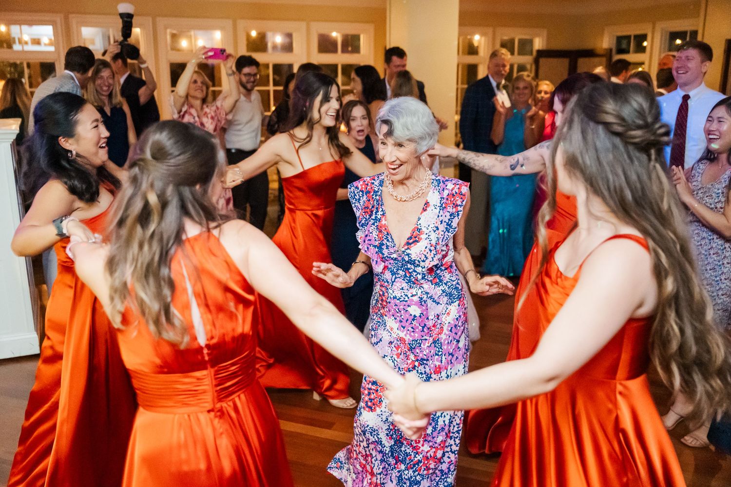 Wedding guests in colorful dresses dancing and celebrating on wooden dance floor at reception.