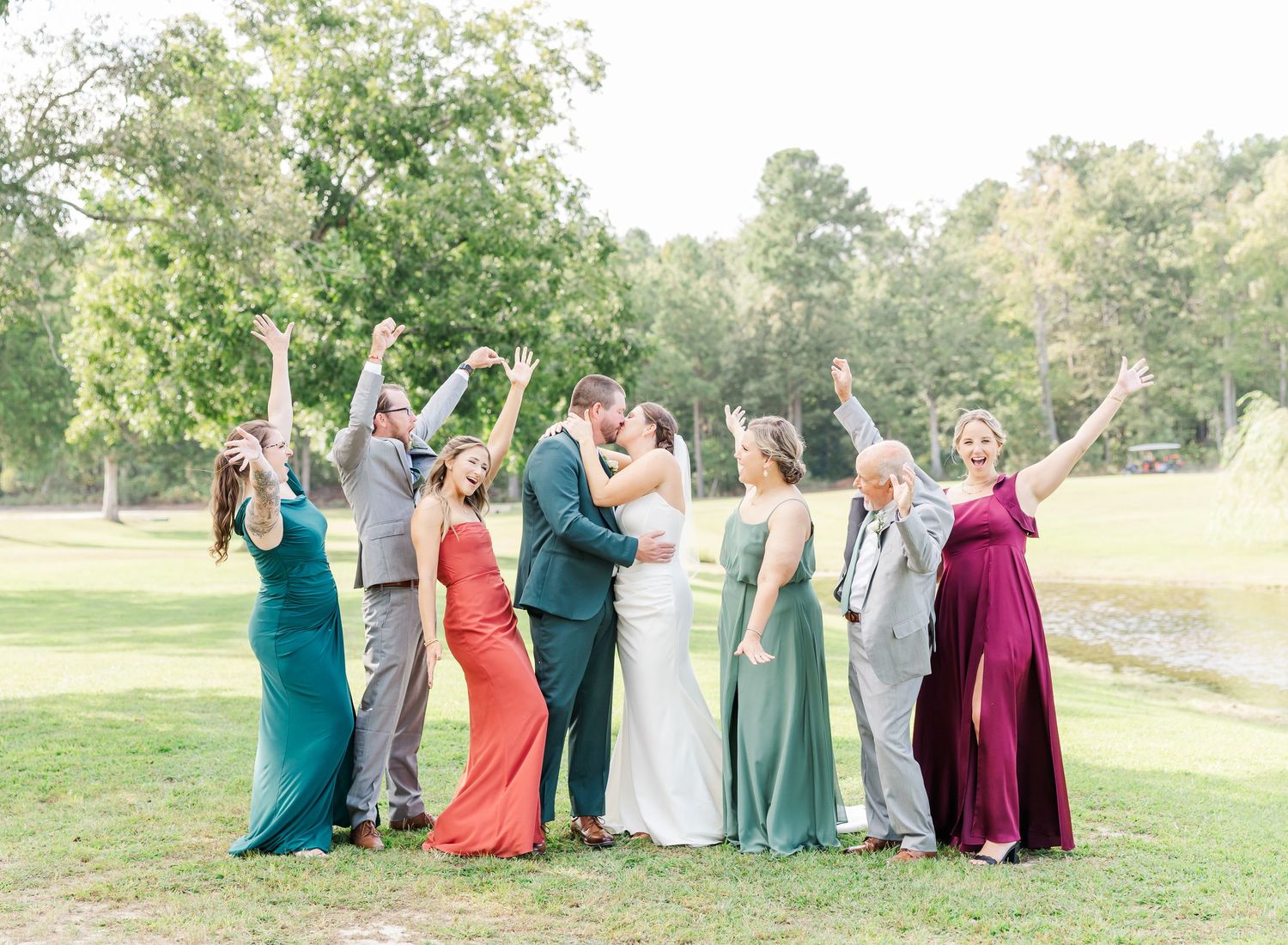 Wedding party in colorful formal attire celebrates joyfully on a sunny lawn with trees in the background.