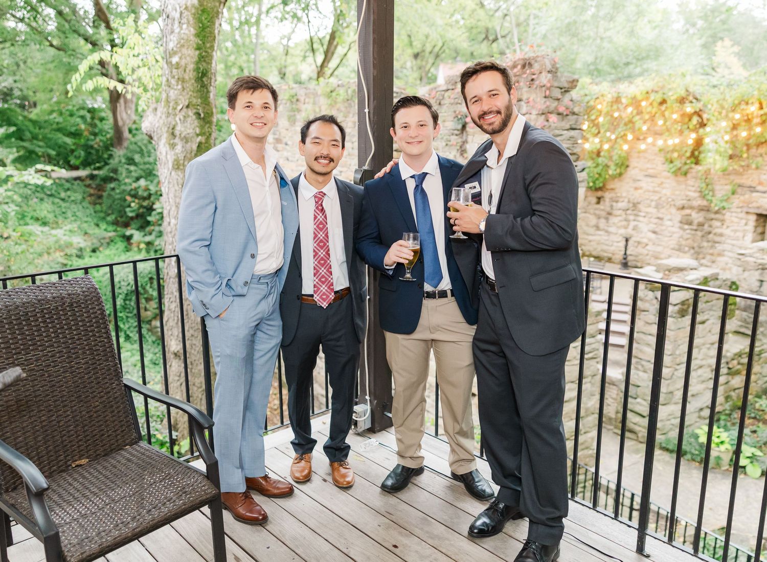 Wedding party members in formal attire gather on an outdoor wooden deck surrounded by greenery.