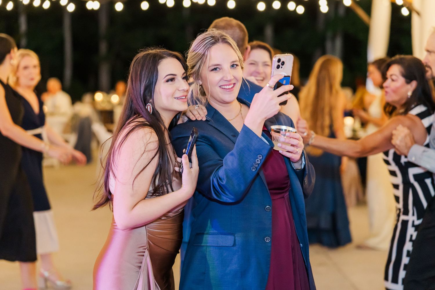 Two women smiling and taking a selfie at an elegant outdoor wedding reception celebration.