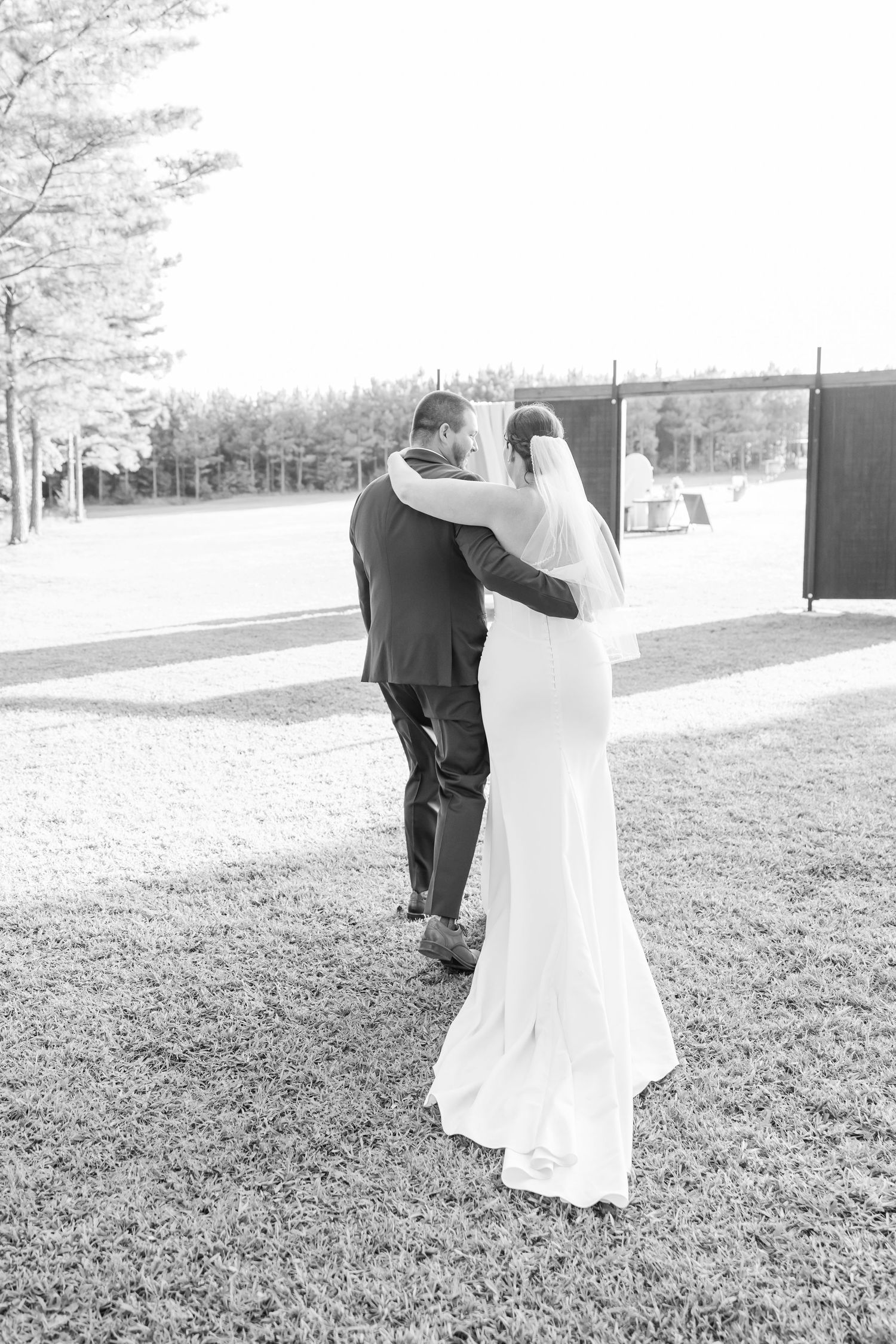 Black and white photo of a bride and groom walking arm in arm across a grassy lawn on their wedding day.