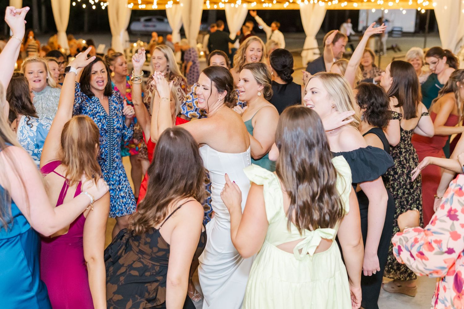 Guests in formal attire dance together on a crowded wedding reception dance floor under warm lighting.