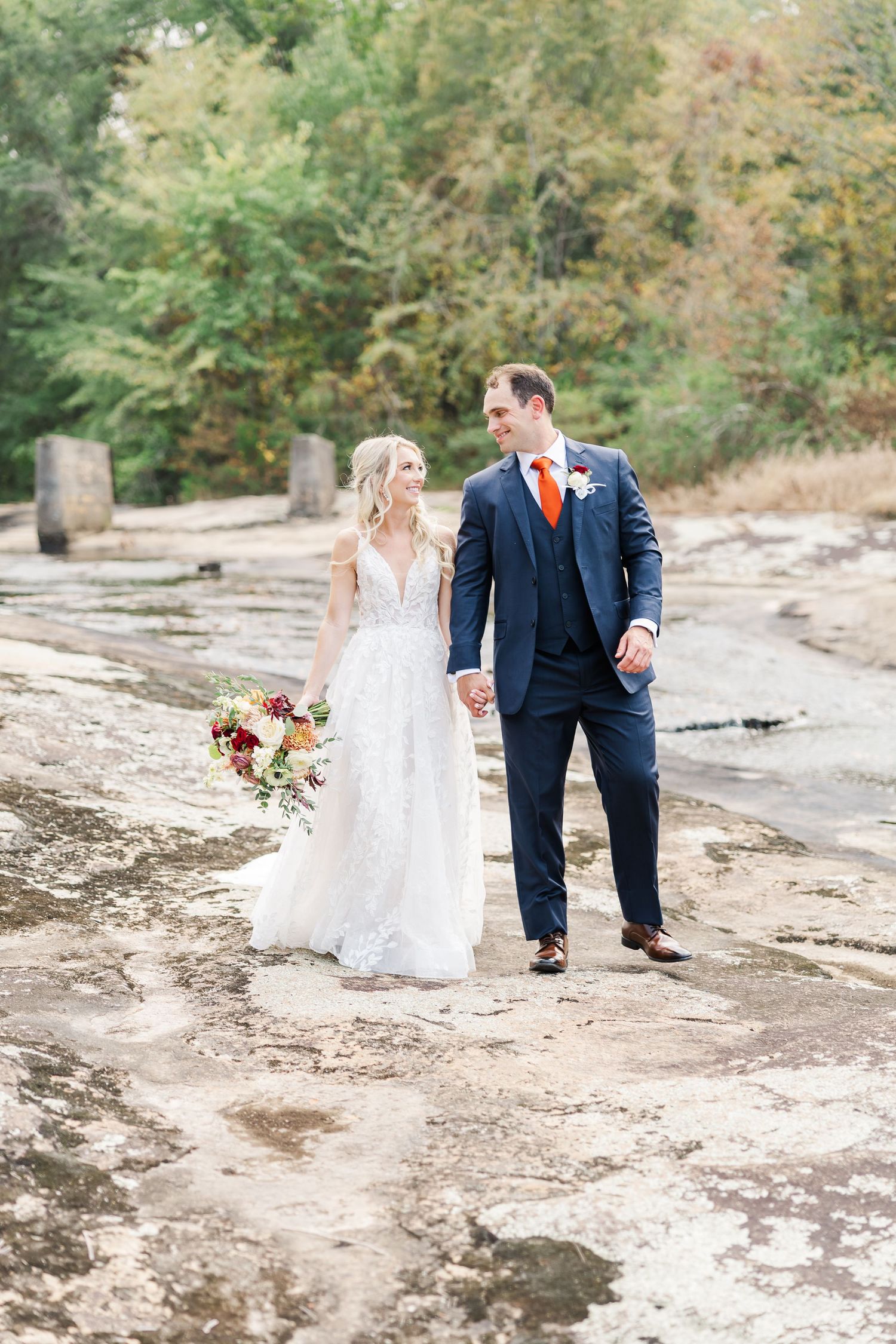 Wedding couple walks along a rocky riverbed surrounded by autumn trees during their outdoor photo session.