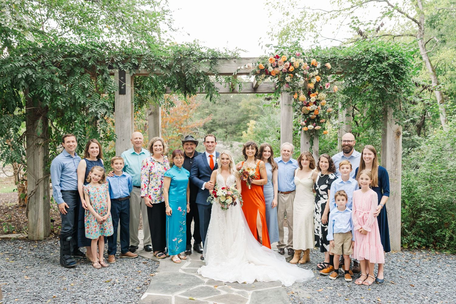 Wedding party poses together under a floral-decorated wooden pergola in an outdoor garden setting.